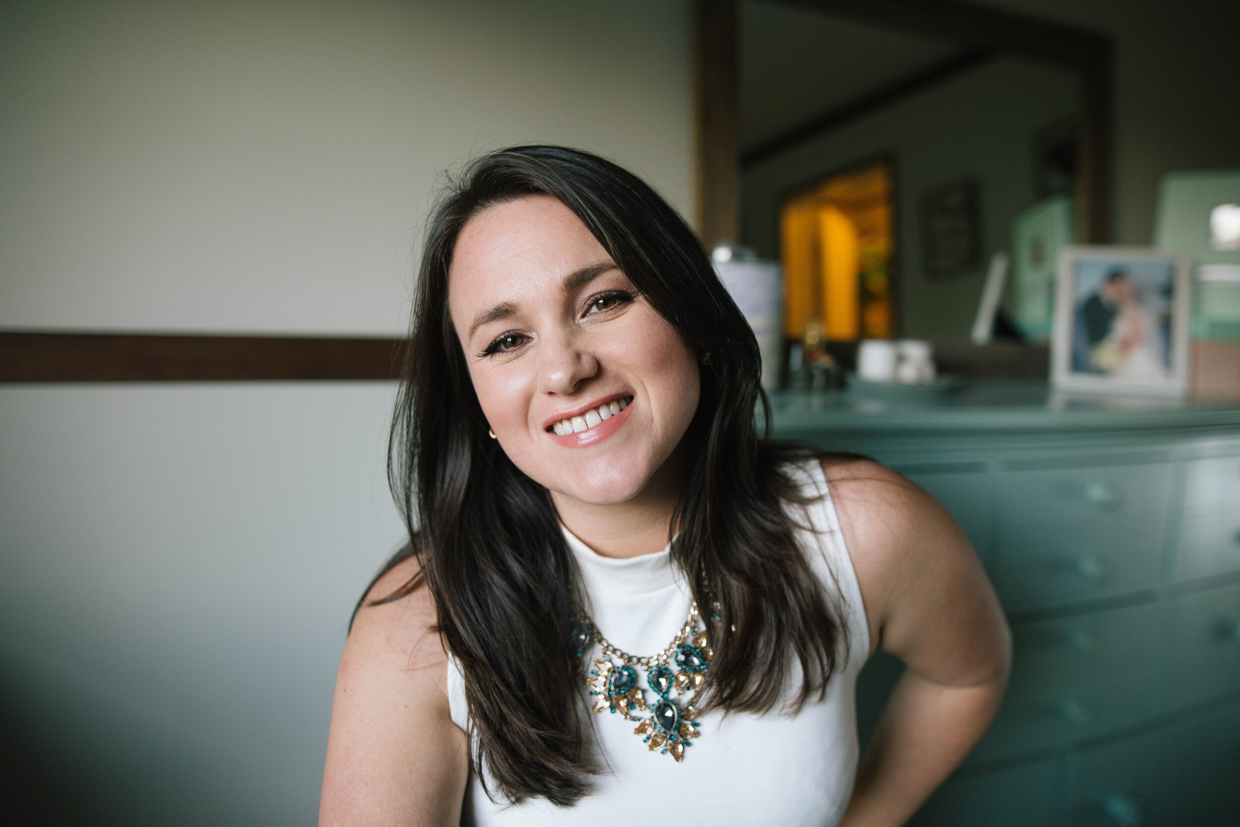 Young woman with dark hair smiling, wearing a white top and a large, blue statement necklace, in a room with framed photographs on a mint green wall.
