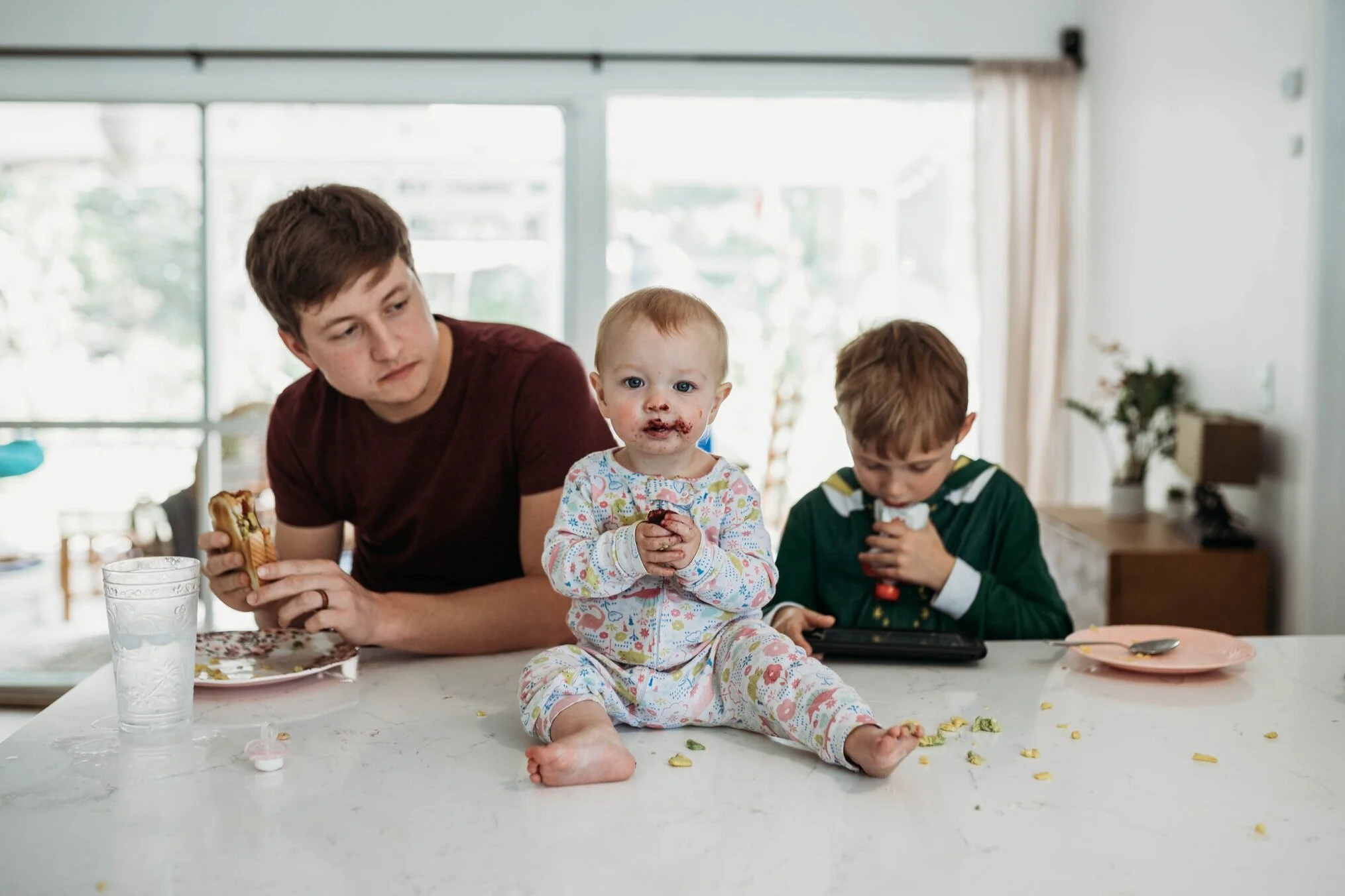A young man and two children are sitting at a table, with messy food and crumbs scattered on the surface. The young man is holding a sandwich, while the girl, who has food on her face, is sitting with her legs extended and eating. The boy is focused 