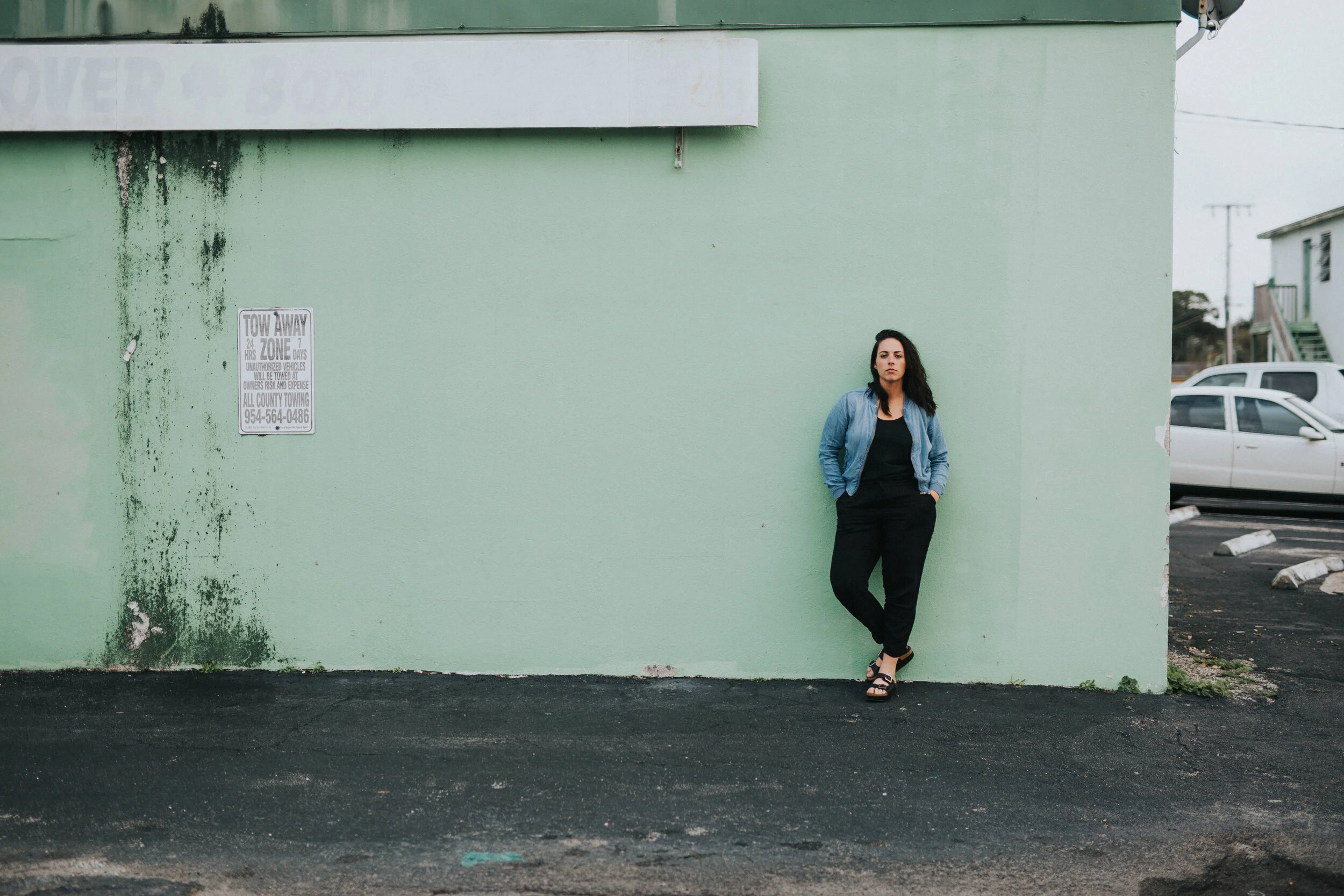 A woman standing against a pastel green building wall. She has dark hair, wearing a blue denim jacket and black pants, with hands in pockets. There is a parked white car and another building in the background.