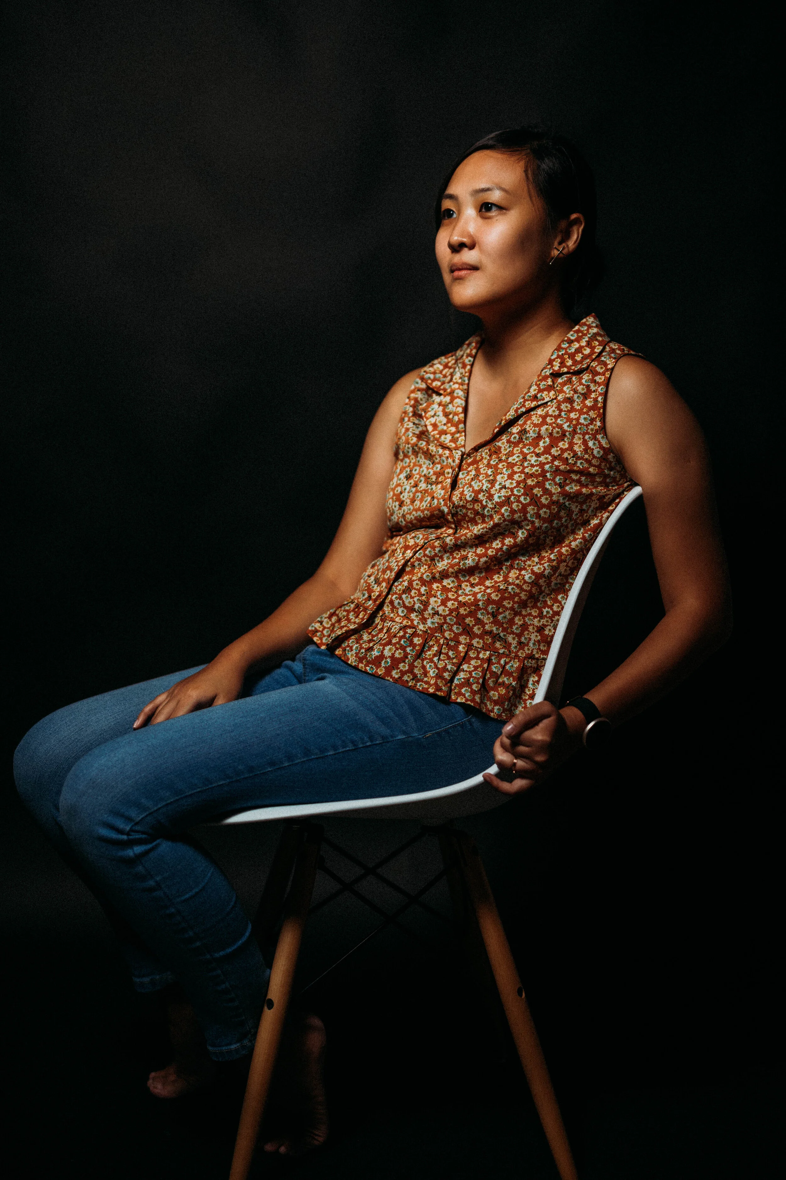 A woman sitting on a white chair against a black background, wearing a sleeveless floral top and blue jeans, with short black hair and minimal makeup.