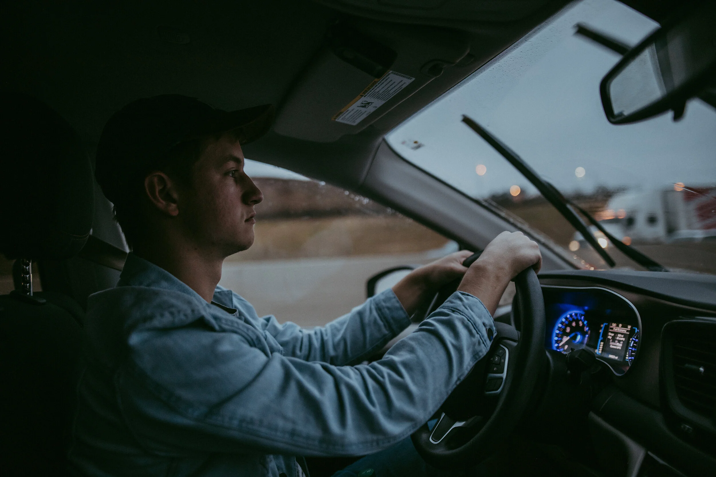 A young man driving a car during a rainy evening, with rain droplets on the windshield and blurred city lights in the background.