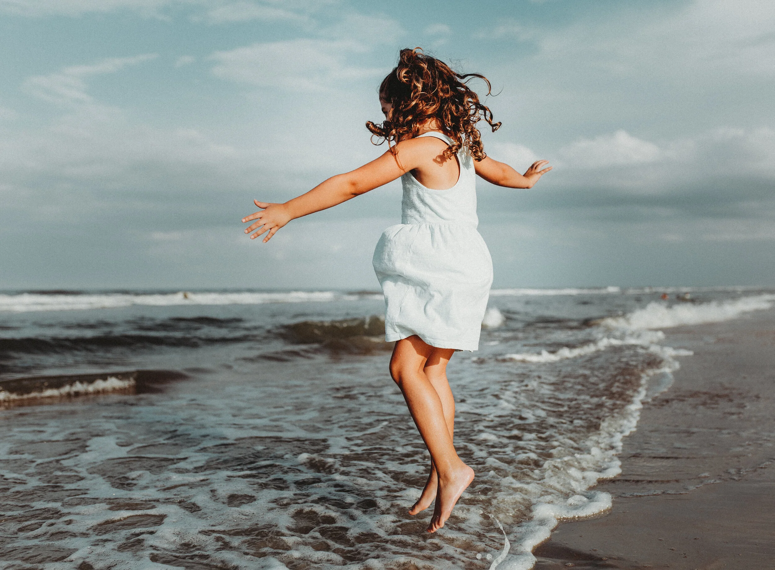 A young girl with curly hair wearing a white dress is walking barefoot on the beach near the water, with her arms outstretched as she faces the ocean. The sky is partly cloudy.