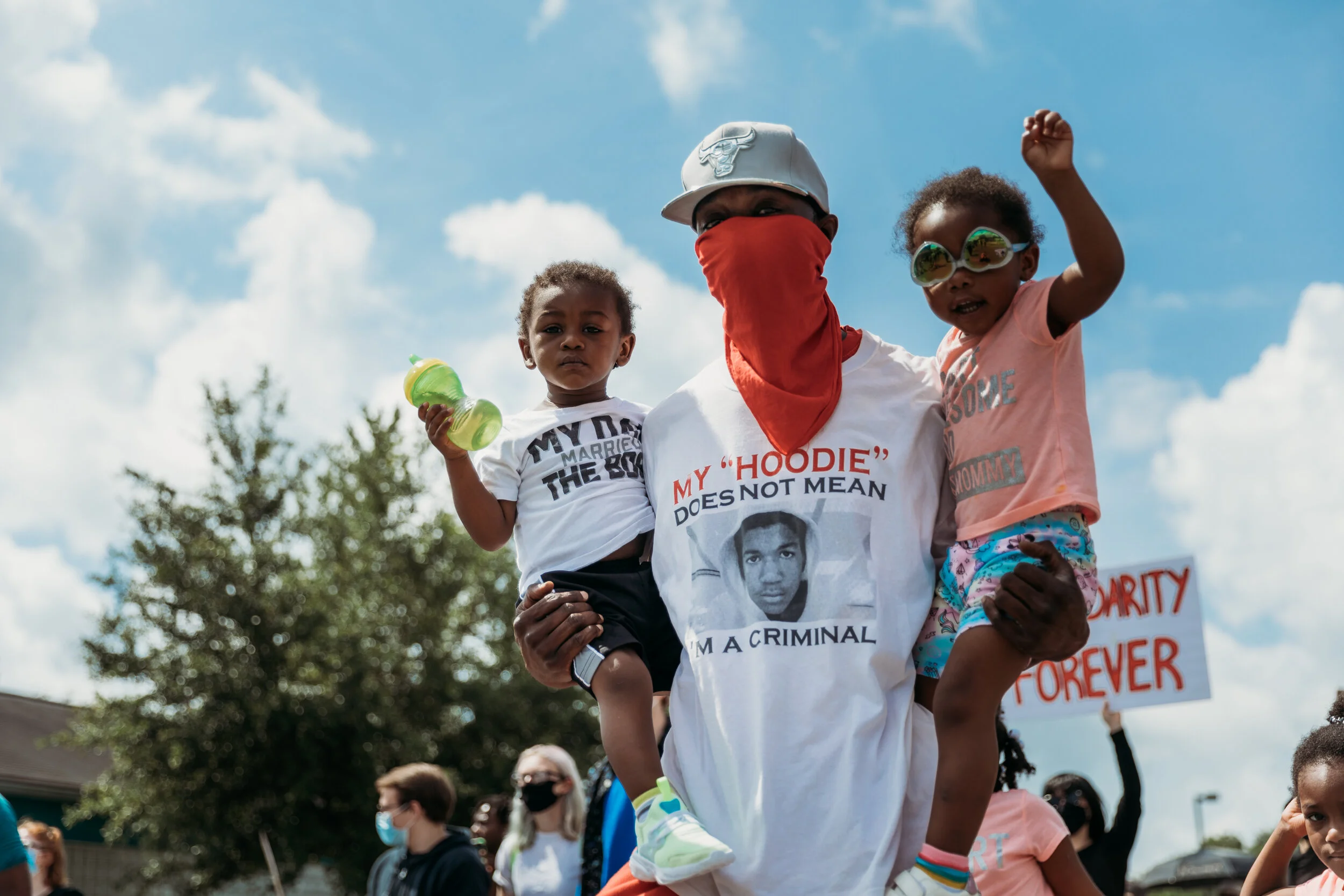 A man wearing a white baseball cap, an orange face mask, and a white T-shirt holding two children on his shoulders during a protest or rally. The children are wearing casual clothes and sunglasses, one of them raising his fist. The background has a b