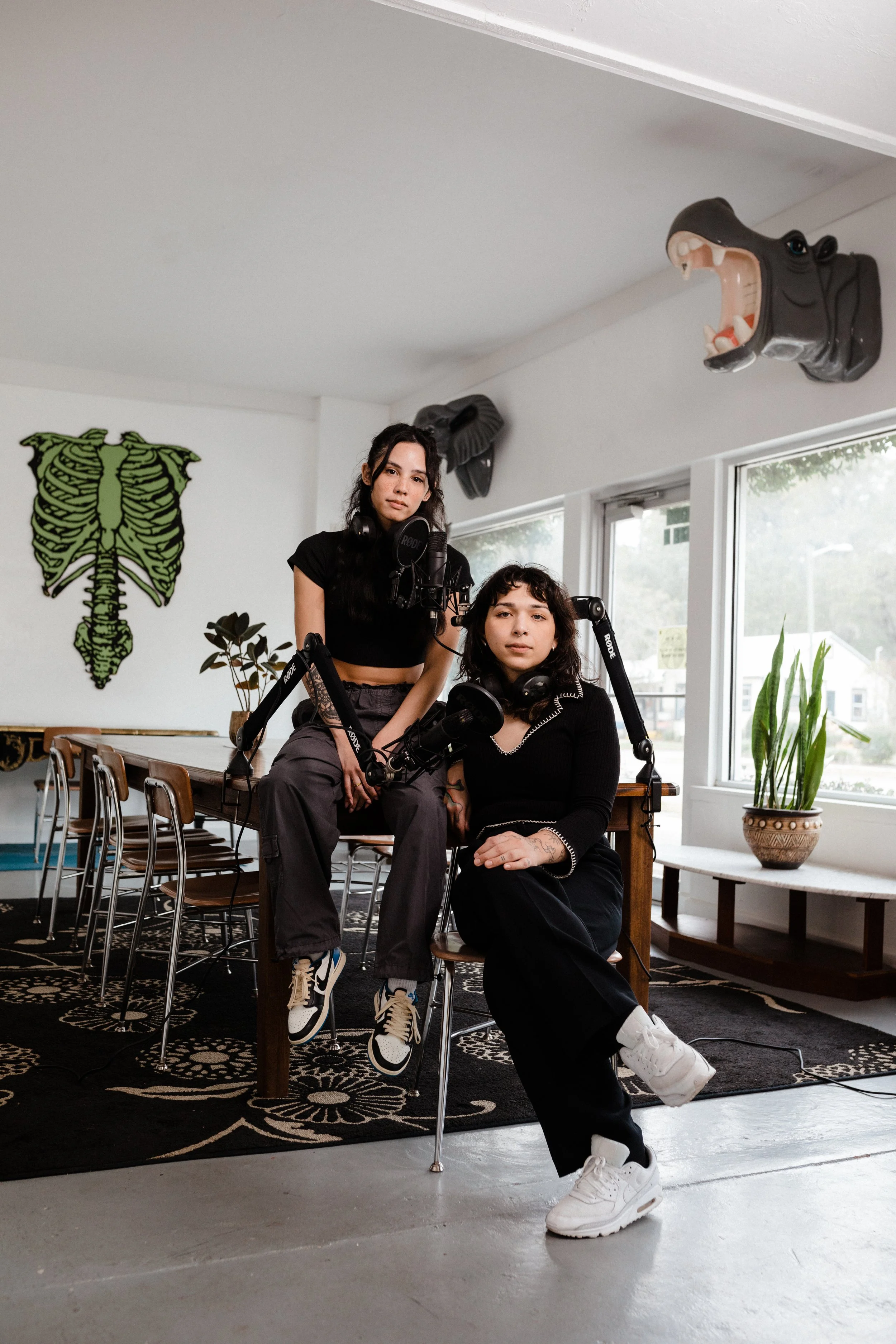 Two women in a room with a black and white patterned rug, a table, chairs, and plants. One woman is sitting and the other is standing. The room has a large window, plants, a wall art of a green skeletal fish, and a decorative hippopotamus head mounte