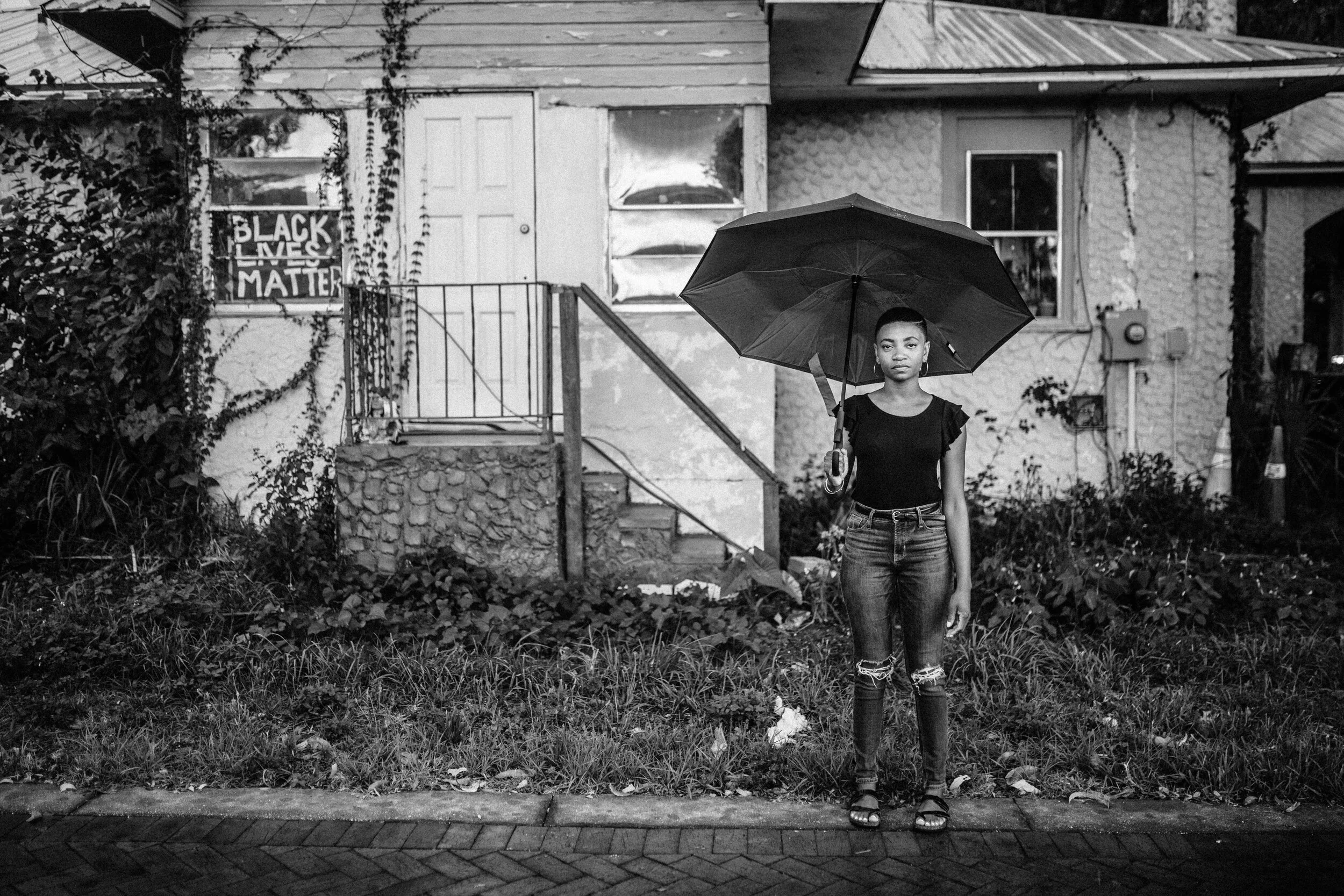 A young woman standing on a sidewalk, holding an umbrella, in front of a house with a 'Black Lives Matter' sign in the window, black and white photo.