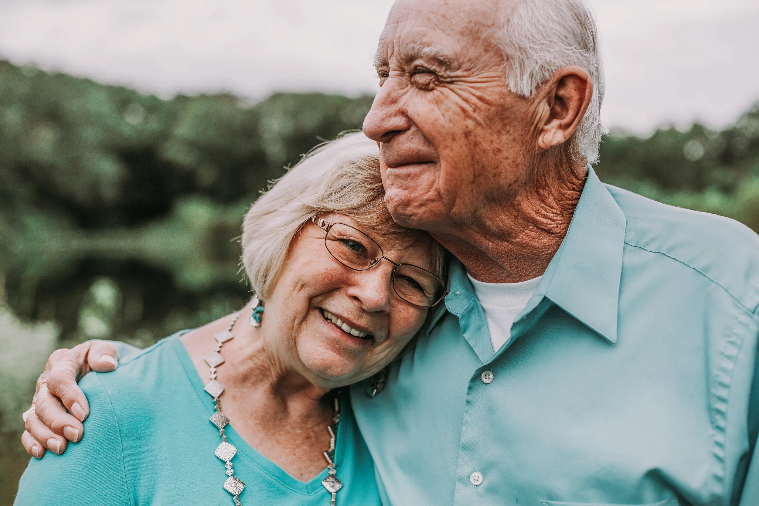 A smiling elderly man and woman hugging outdoors, blurred green background.
