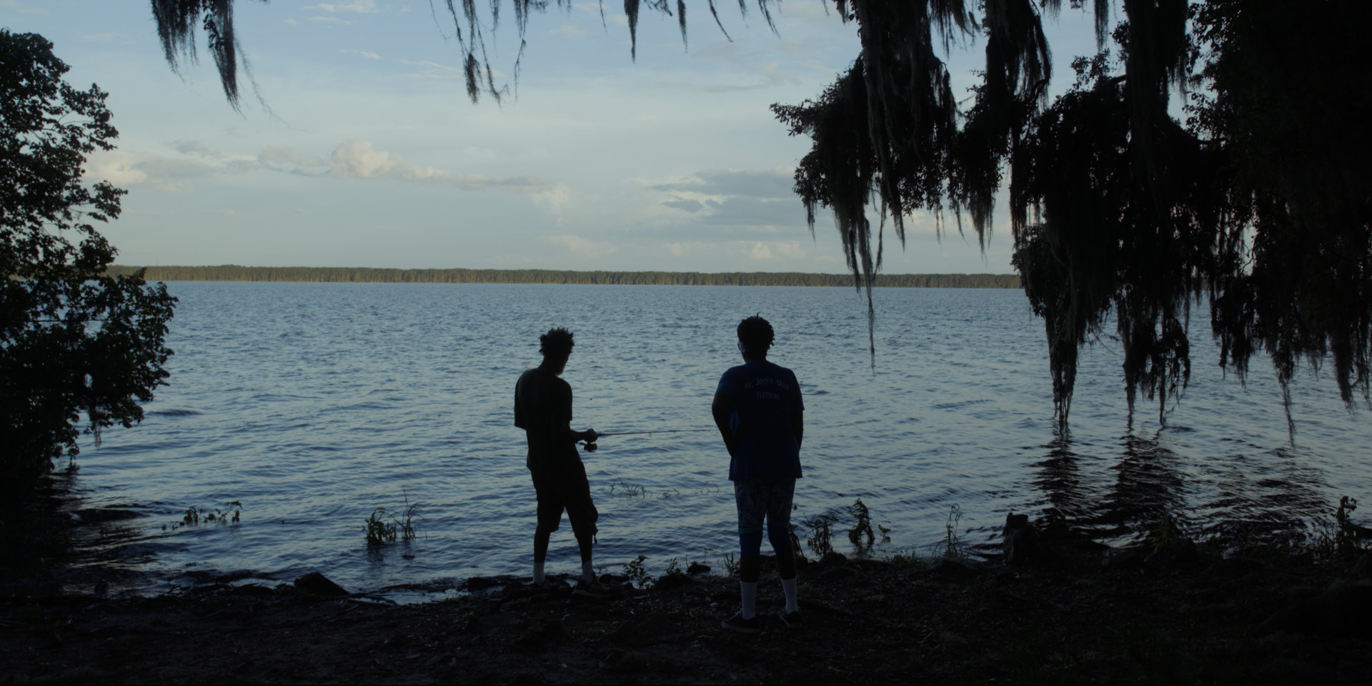 Two people stand by a lake under hanging moss, one holding a fishing pole, as the sun sets or rises in the distance over the water.