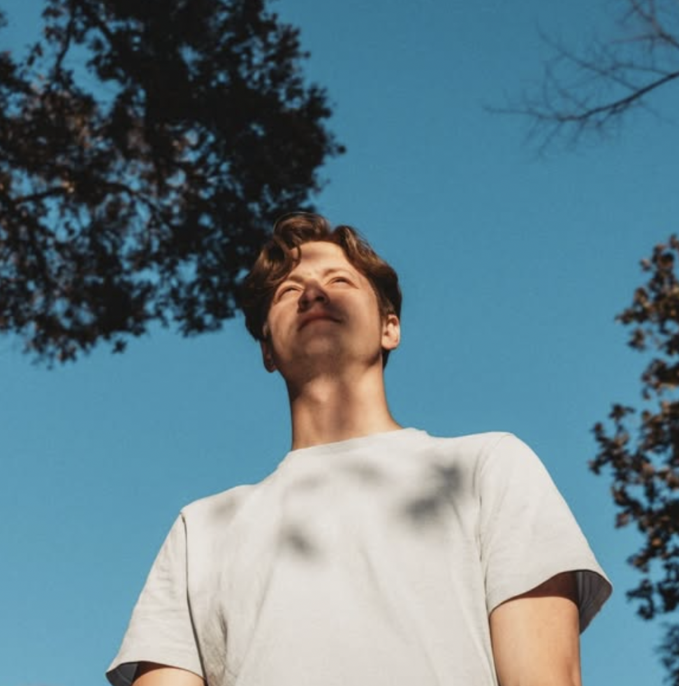 A young man standing outdoors looking up at the sky, with trees around him and a clear blue sky in the background.