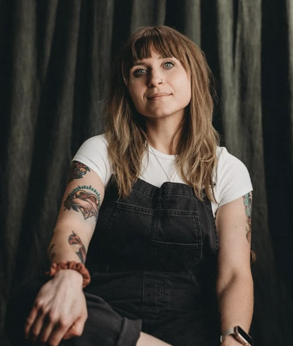 A woman with shoulder-length, wavy brown hair, blue eyes, wearing a white t-shirt with black overalls and tattoos on her arms, standing in front of dark curtains.