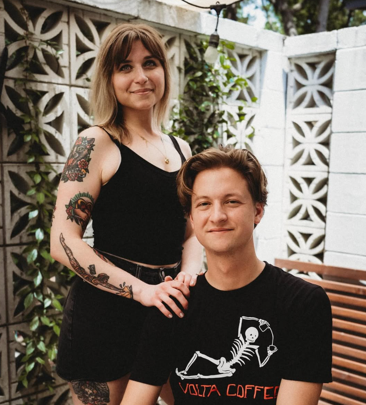 A woman with shoulder-length light brown hair, wearing a black sleeveless top, standing outdoors with a man in front of her. The woman has tattoos on her arms and is holding the man’s hand. The man has short brown hair, is smiling, and wearing a black T-shirt with a skeleton holding a wine glass and the words "Volta Coffee". They are in a patio area with white decorative concrete blocks and greenery in the background.