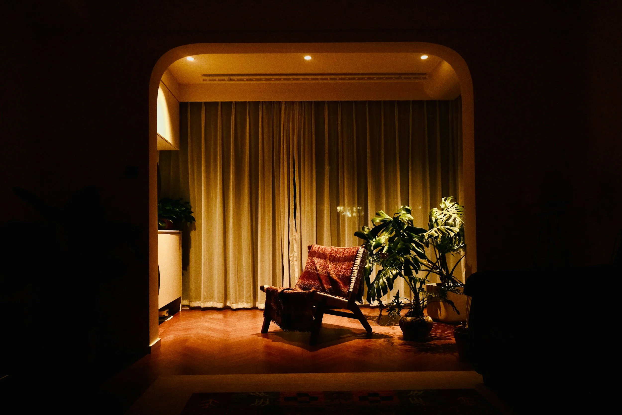 A warmly lit, cozy living room with a chair covered with a red blanket, and several potted plants near large window with closed curtains.