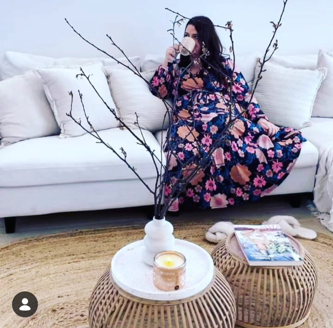 A woman in a floral dress sitting on a white couch, drinking from a glass mug, with a leafless branch arrangement, a candle, and magazines on rattan tables in front of her.