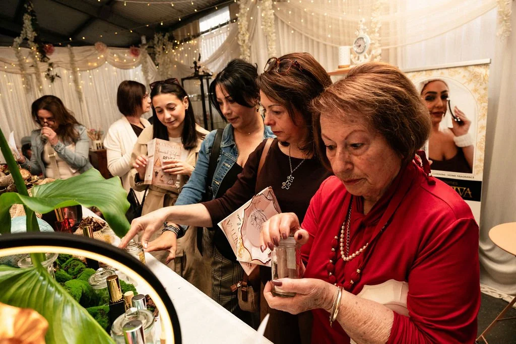 Group of women at a craft or beauty event, examining products on a table decorated with green plants, in a decorated, festive environment with string lights and draped fabric. Scentsetiva Elemental Perfume reveal launch with people loving the aromas.