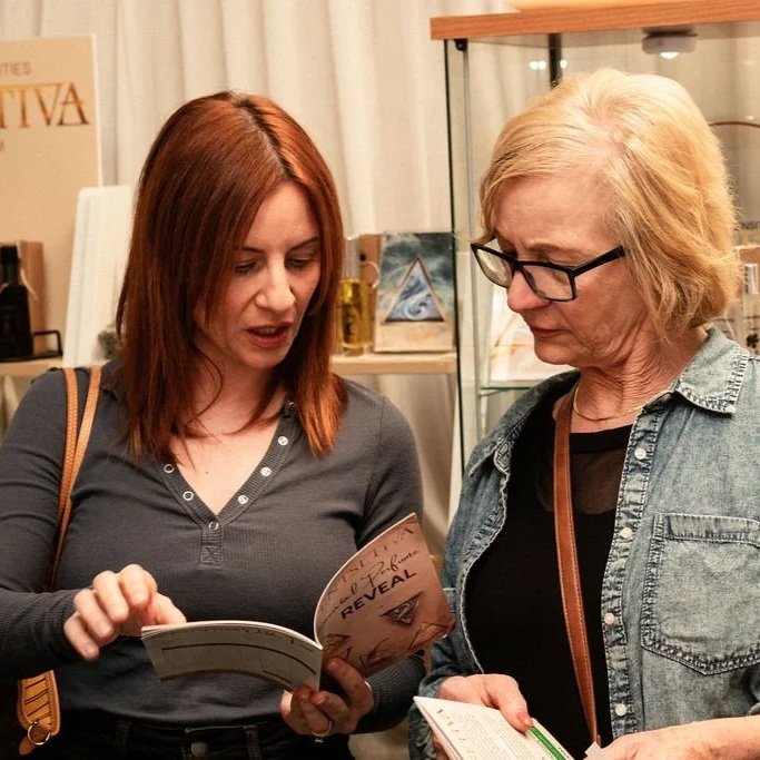 Two women standing together, reading a book or brochure at a Scentsetiva elemental perfume reveal browsing our catalogue