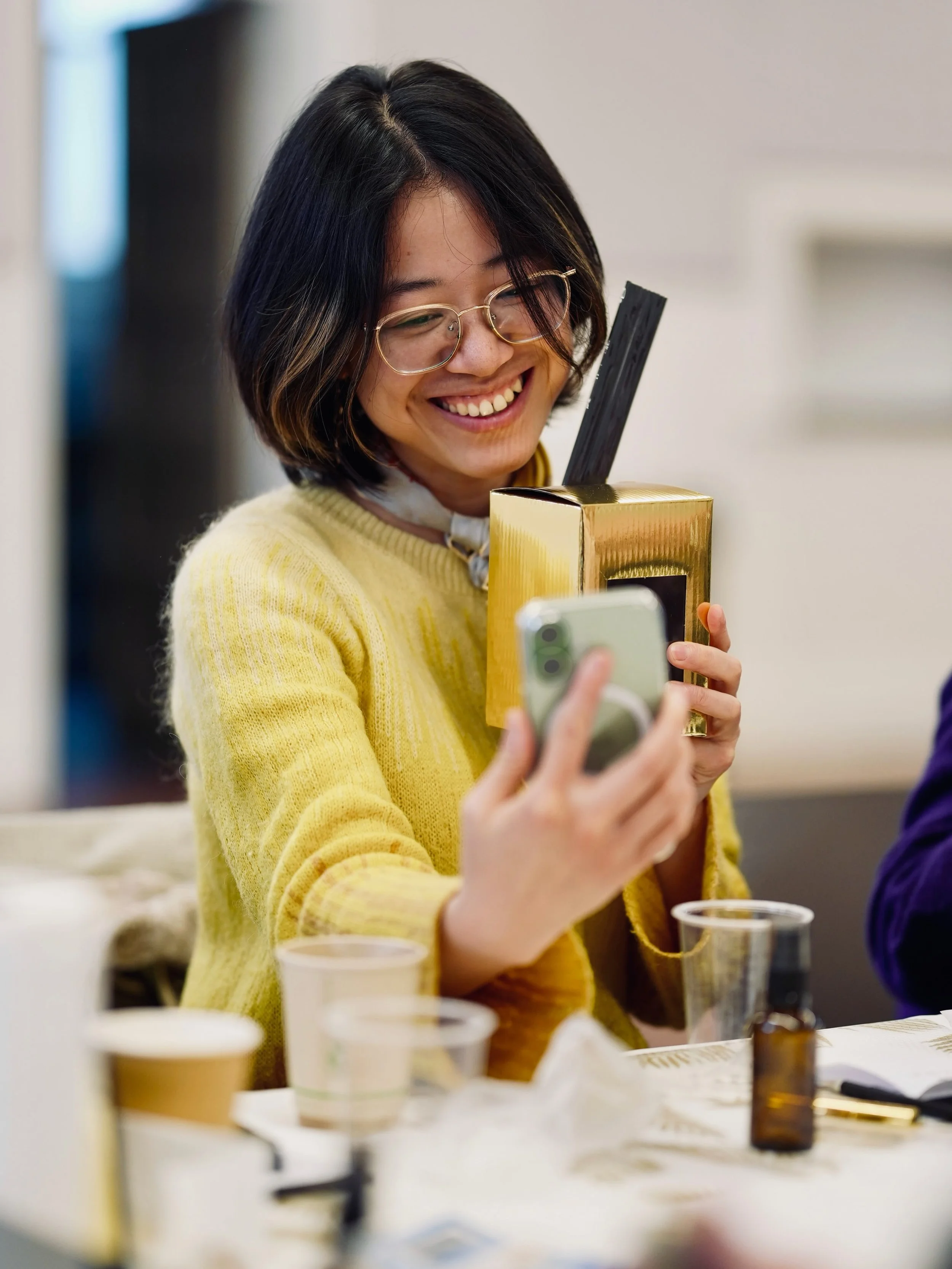 Woman with short dark hair, glasses, and a yellow sweater smiling and taking a photo with her phone during a social gathering.