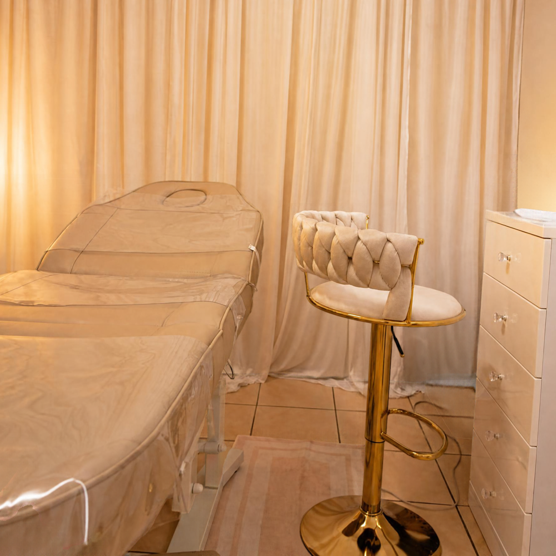 A beige, padded, velvet armchair with gold trim and a cream-colored, tufted backrest, positioned next to a gold-colored pedestal table in a room with beige curtains and a white dresser.