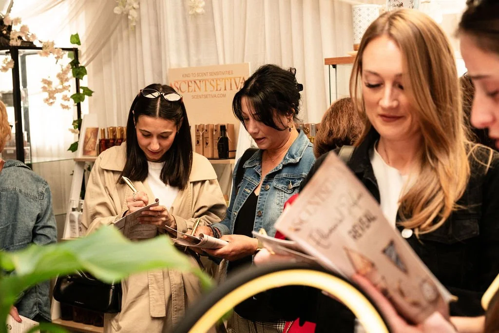 Group of women at Scentsetiva Elemental Perfume Reveal browsing at a launch  or trialling perfumes at the event, holding pamphlets and elemental quiz guide magazines, in an indoor setting with display shelves and a scentsetiva sign in the background.