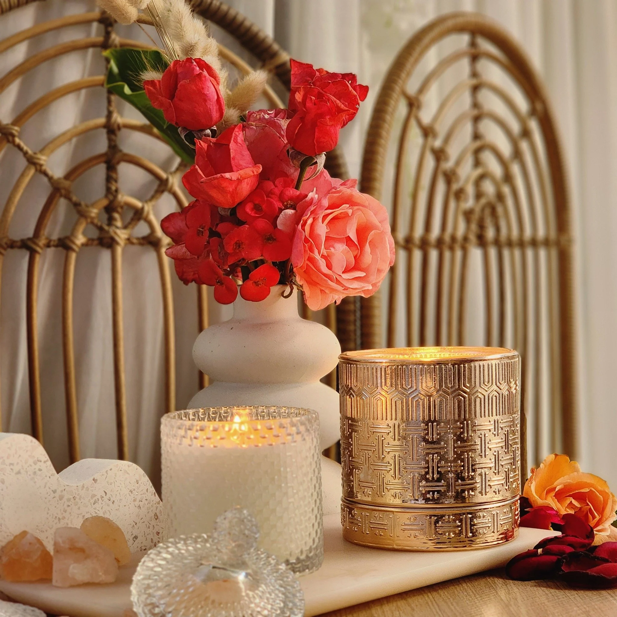 Vase with a bouquet of red and pink flowers on a table with candles, gold decorative container, and assorted sweets.