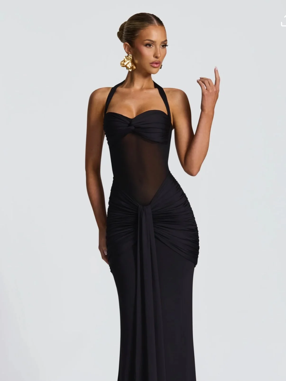 A woman wearing a black evening gown with a sheer midsection and gold earrings, posing against a plain light gray background.