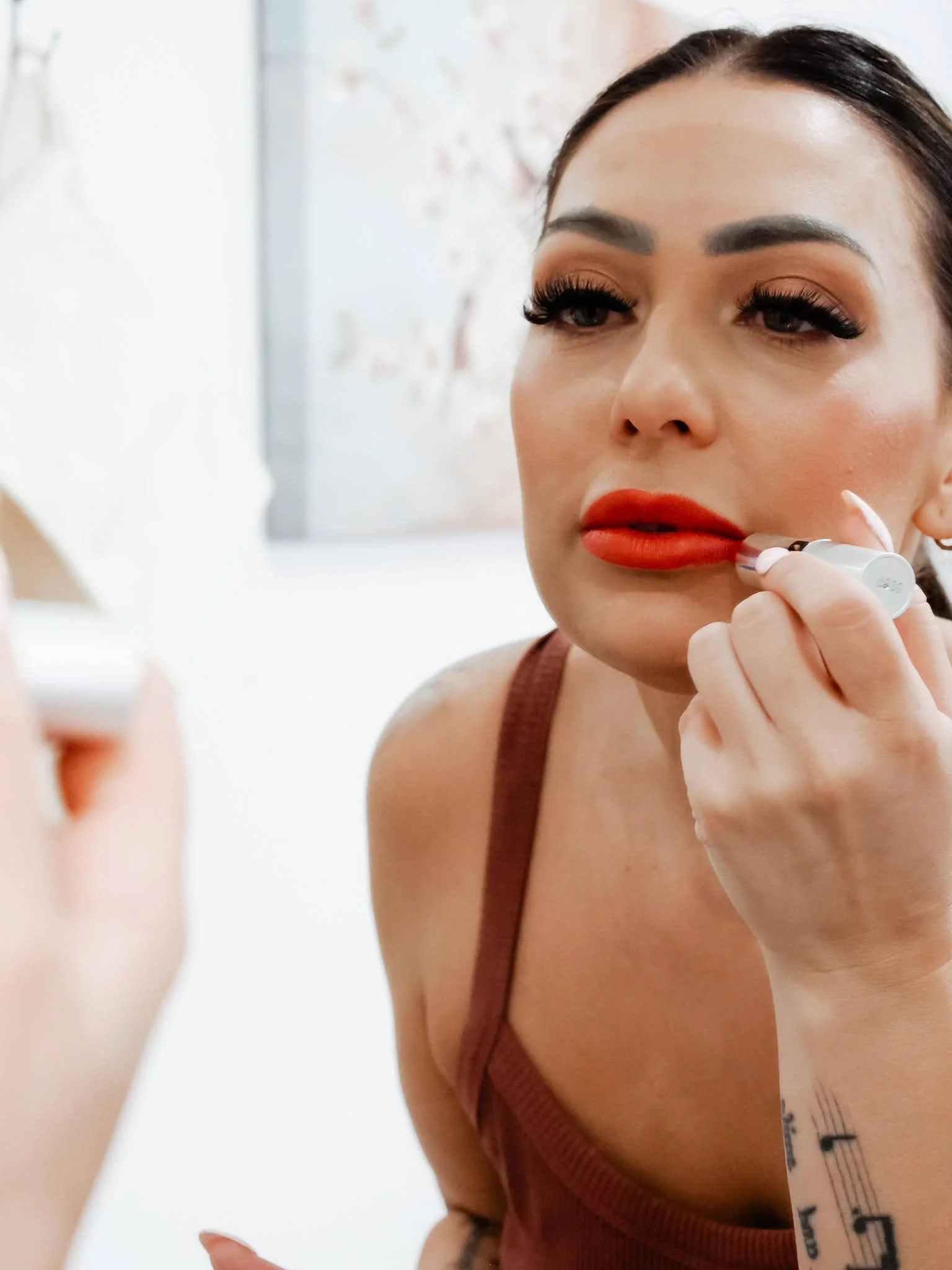 A woman applying lipstick in front of a mirror, holding a white lipstick tube and wearing a brown tank top.
