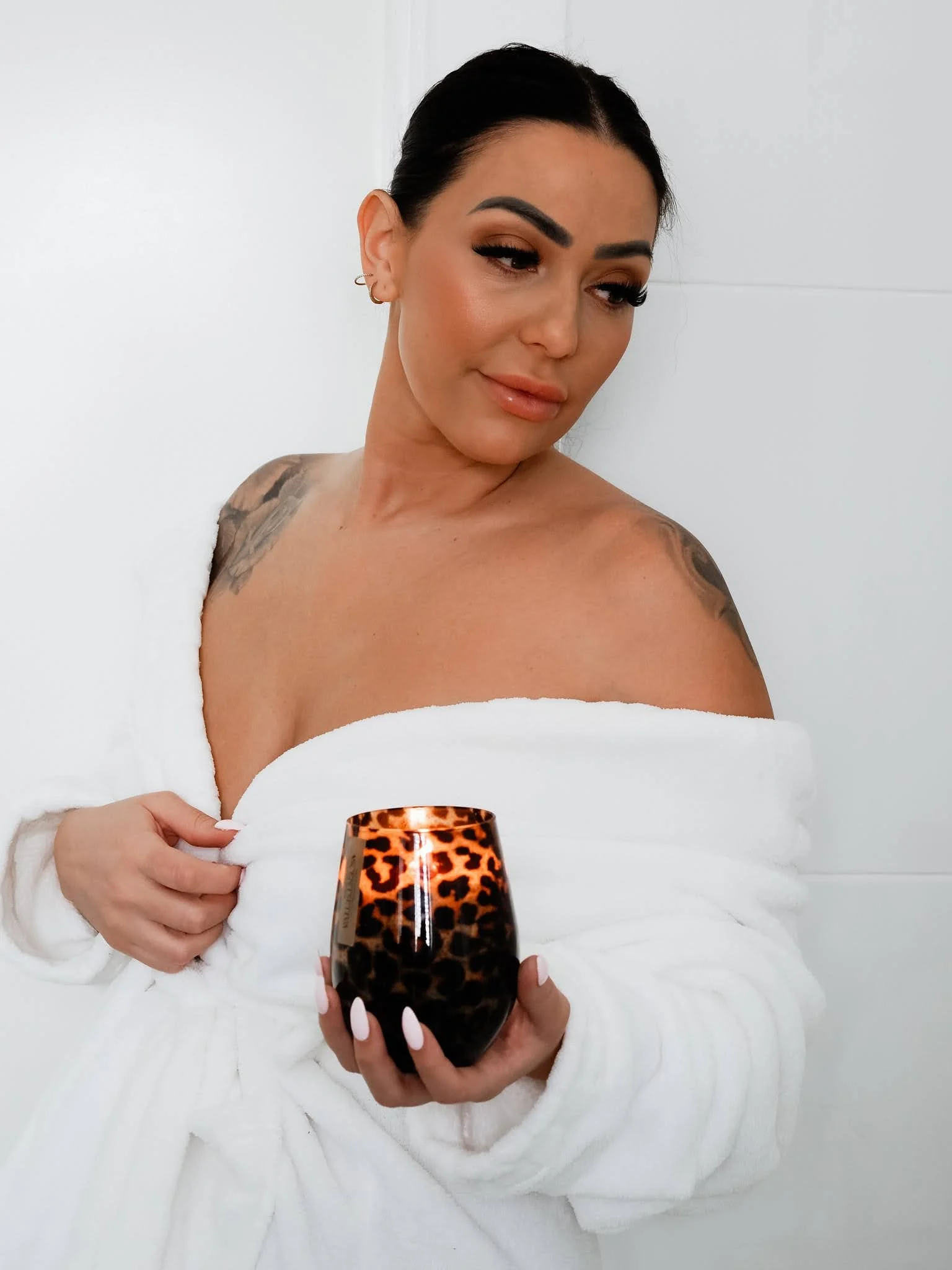 A woman with dark hair with Scentsetiva Candle, wrapped in a white towel, holding a leopard print wine glass in front of her, against a white wall background. Ready for a bath with Scentsetiva Velvet Glow Body Wash and Velvet Glow Body Moisturiser