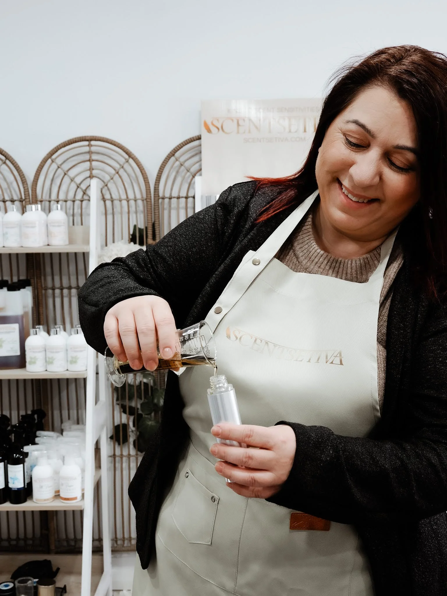 A woman with dark hair and a light-colored apron pouring a liquid from a test tube into a small bottle in a room with shelves of bottles in the background.
