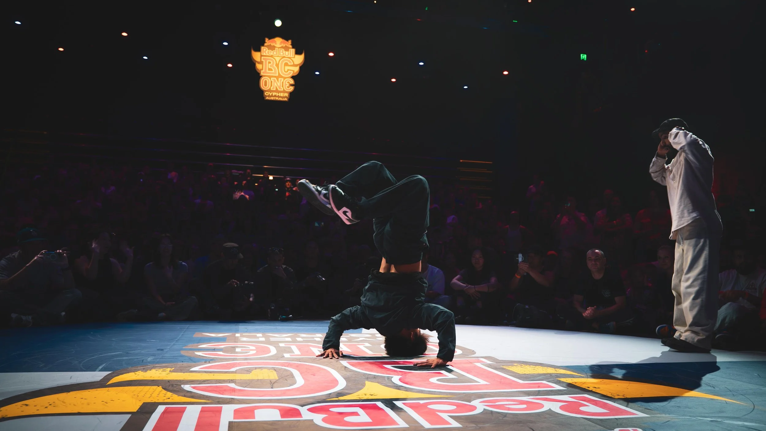Un ballerino in piedi fa uno stand in equilibrio su una mano sul palco del Red Bull BC One, circondato da un pubblico e illuminato da luci sparse sopra. Il logo del evento è visibile sullo sfondo.