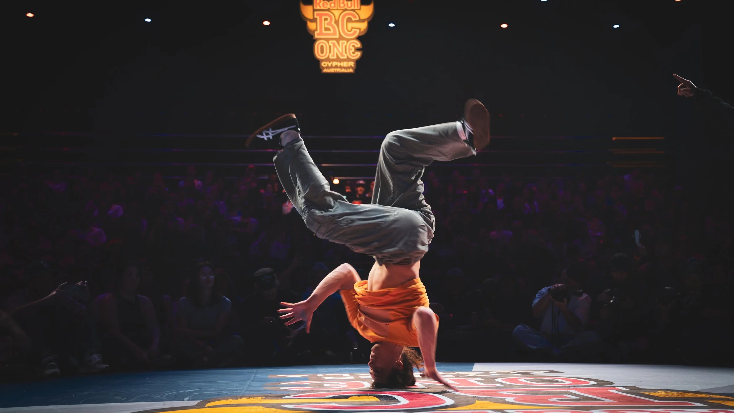 Un ballerino che esegue un colpo di breakdance durante una competizione, con un pubblico seduto al buio e un logo di Red Bull BC One sullo sfondo.