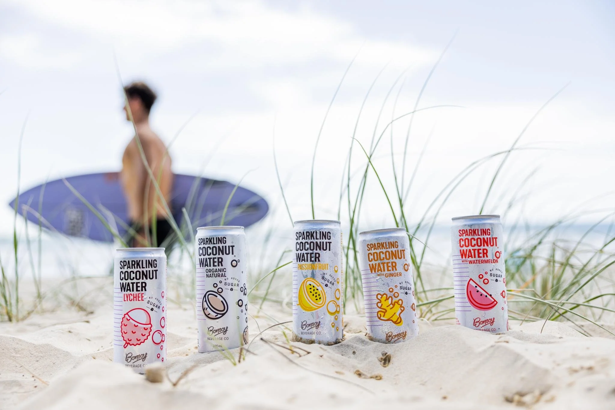Bottiglie di acqua frizzante aromatizzata al cocco e altri frutti posizionate sull'acqua sulla sabbia con un ombrellone e una persona sfocata in spiaggia