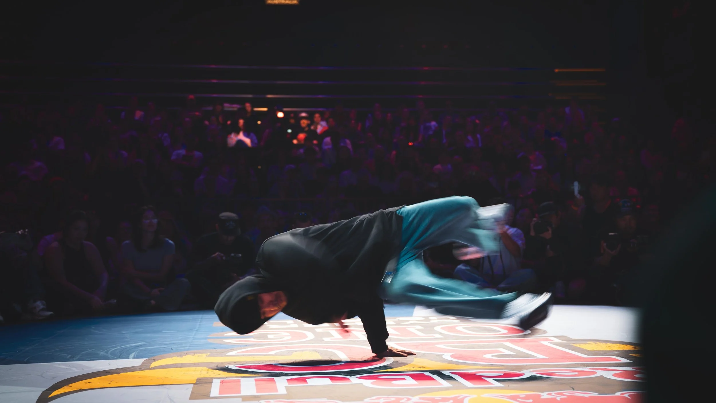 Un ballerino sta eseguendo una mossa di breakdance sul pavimento di un'arena affollata, con un pubblico seduto che lo guarda. L'ambiente è scuro con luci soffuse, e il ballerino si veste con abiti sportivi di colore scuro.