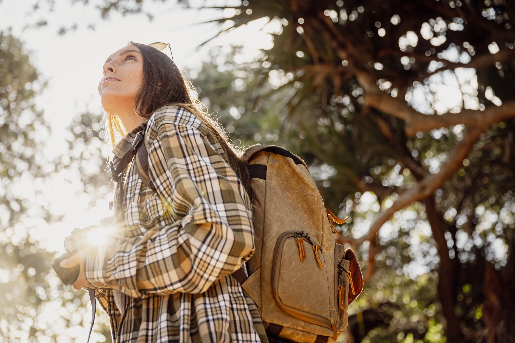 Ragazza con cappello da sole e zaino in spalla al tramonto tra gli alberi, vestita con camicia a quadri.