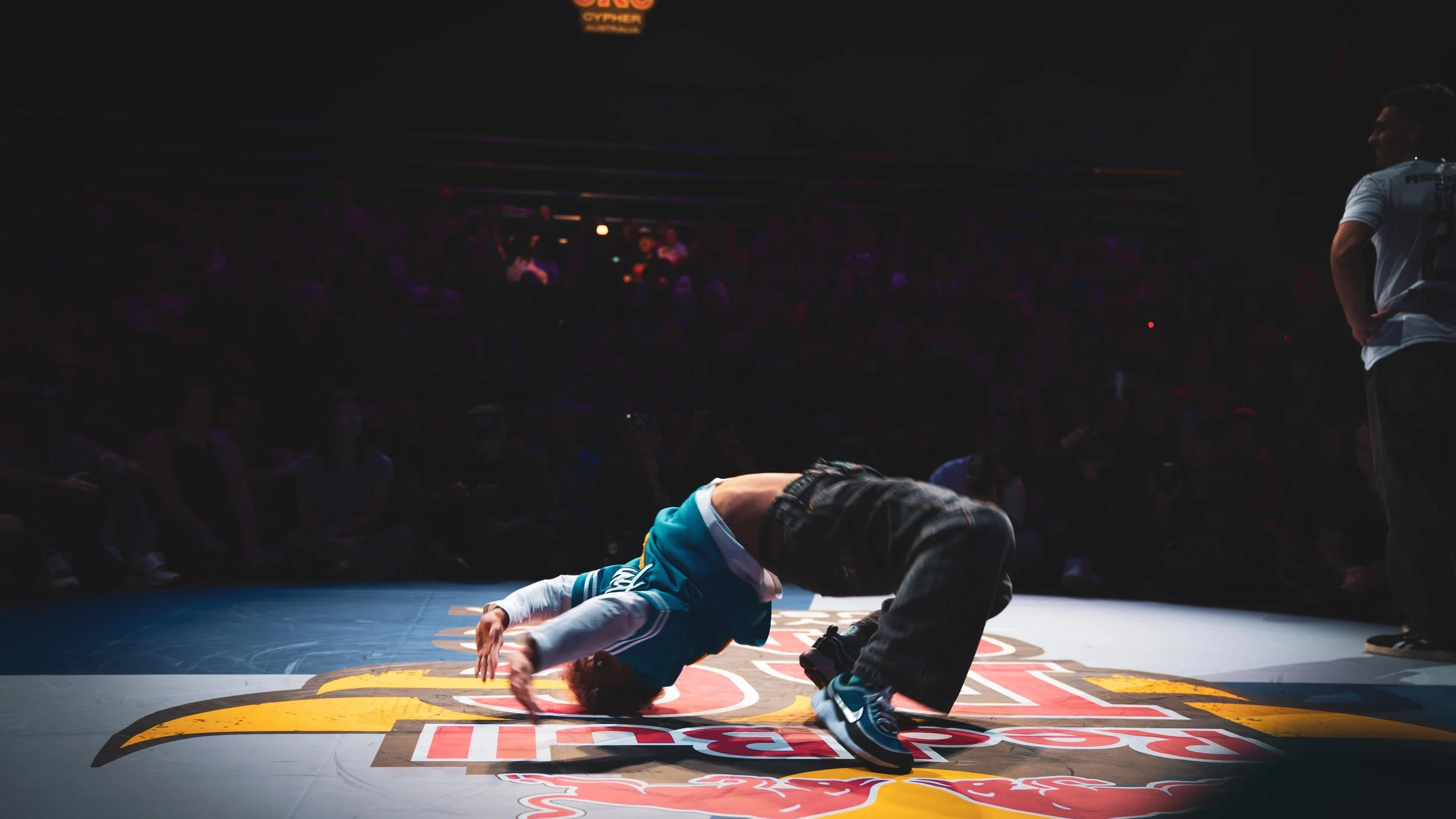 Rissa che si trova a terra sul ring durante una competizione di breakdance, con un giudice o spettatore che osserva in piedi accanto.