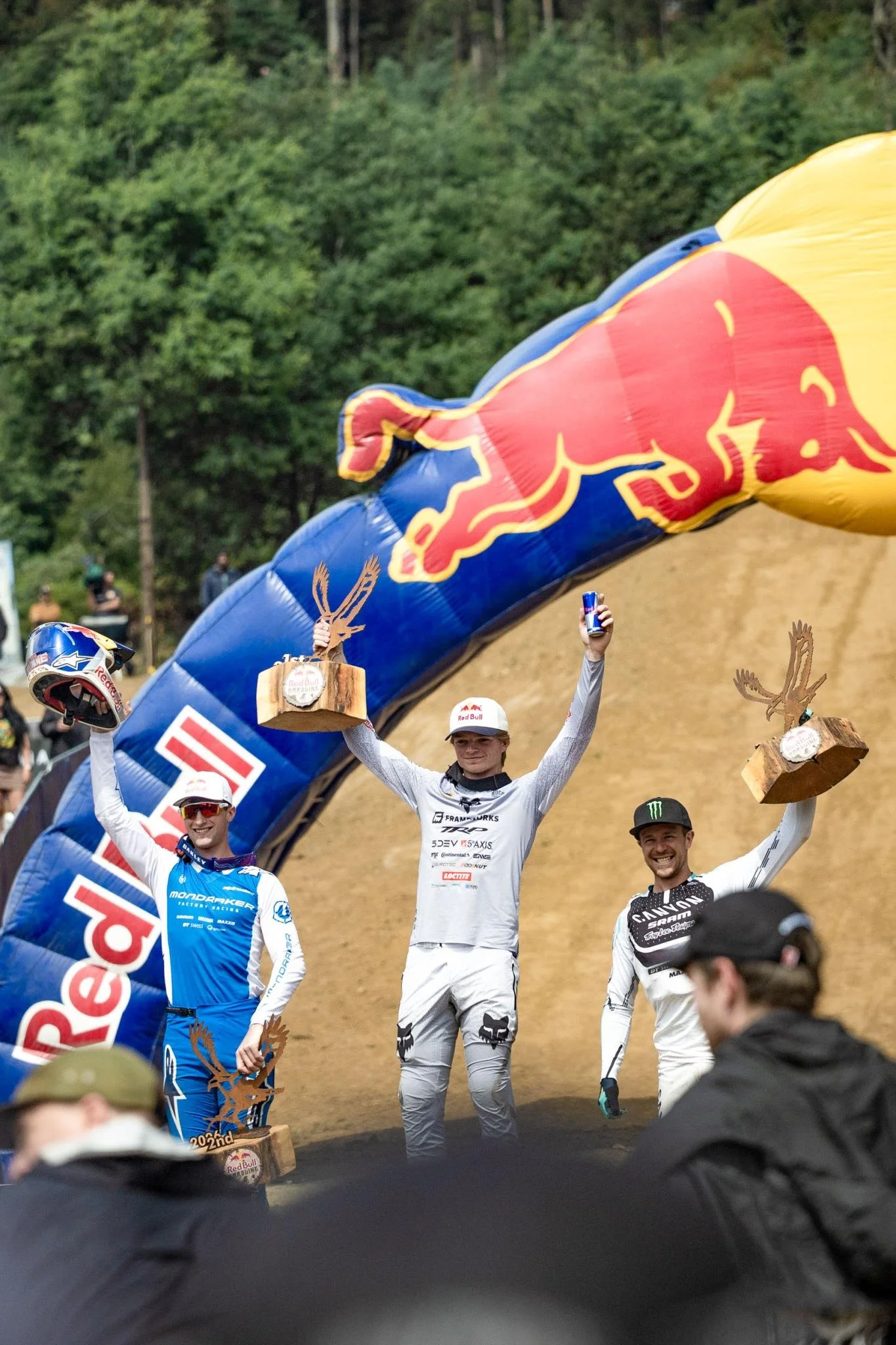 Vincitori di una gara di downhill in Tasmania che festeggiano con trofei e un arco pubblicitario Red Bull sullo sfondo, in un ambiente naturale con alberi.