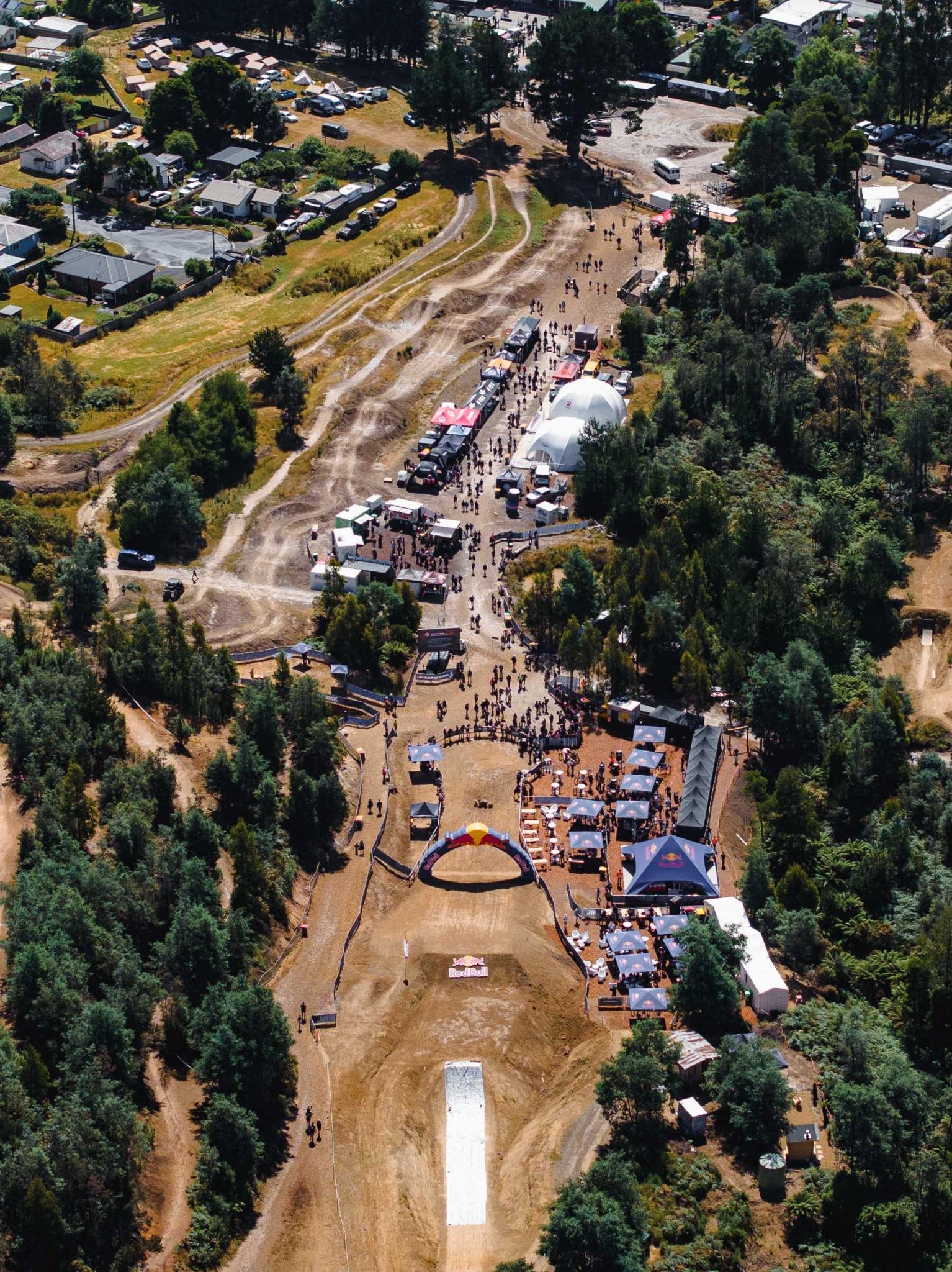 Immagine aerea di un evento all'aperto in un'area boschiva, con molte tende, gazebo e persone che partecipano a varie attività, tra cui una pista di BMX e uno spazio per spettacoli o intrattenimento.