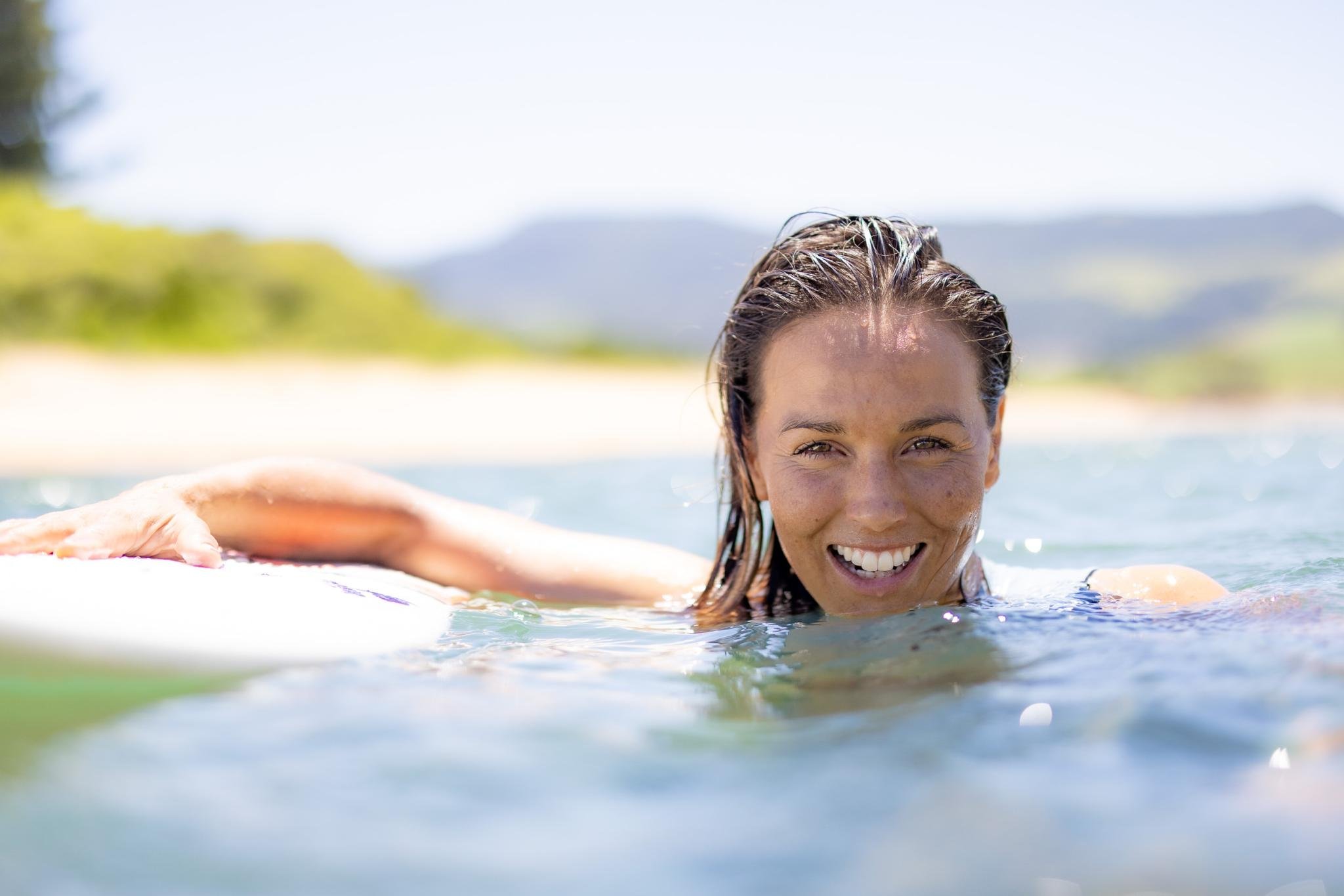 Donna sorridente in mare con la sua tavola da surf difianco.