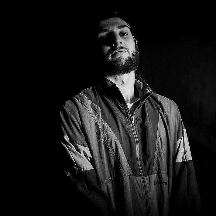 Ritratto in bianco e nero di un uomo con barba, in piedi con le braccia incrociate, indossa una giacca sportiva e guarda verso l'obiettivo.