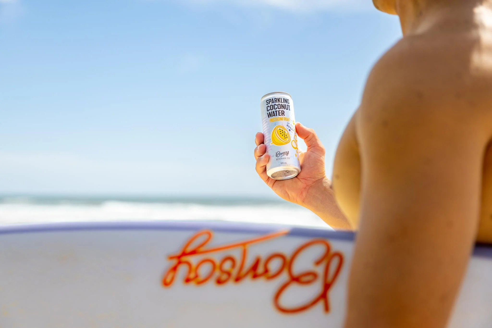 Mano che tiene una lattina di acqua di cocco frizzante al limone e passionfruit sulla spiaggia con il mare sullo sfondo.