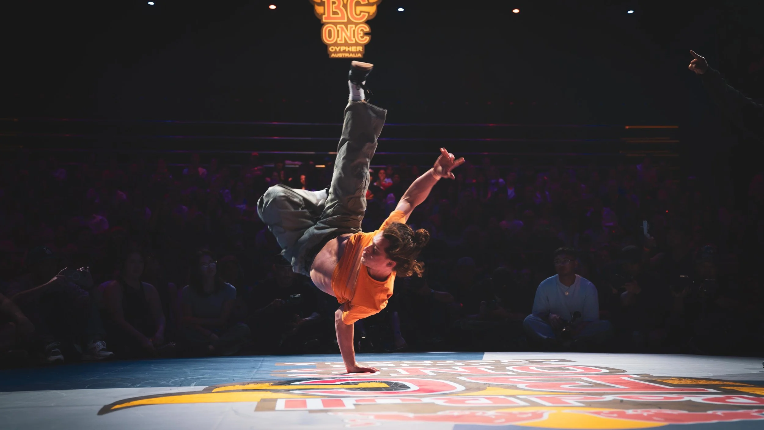 Performer di breakdance che esegue un airchair: freeze su un braccio, appoggiandosi con il gomito sul fianco del corpo. Ballerino presenta un pantalone cargo e una maglietta arancione, sta facendo la competizione Red Bull Bc One Cypher Australia
