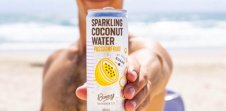 Una persona che tiene in mano una lattina di acqua frizzante al gusto di cocco e frutto della passione, con una spiaggia sullo sfondo.