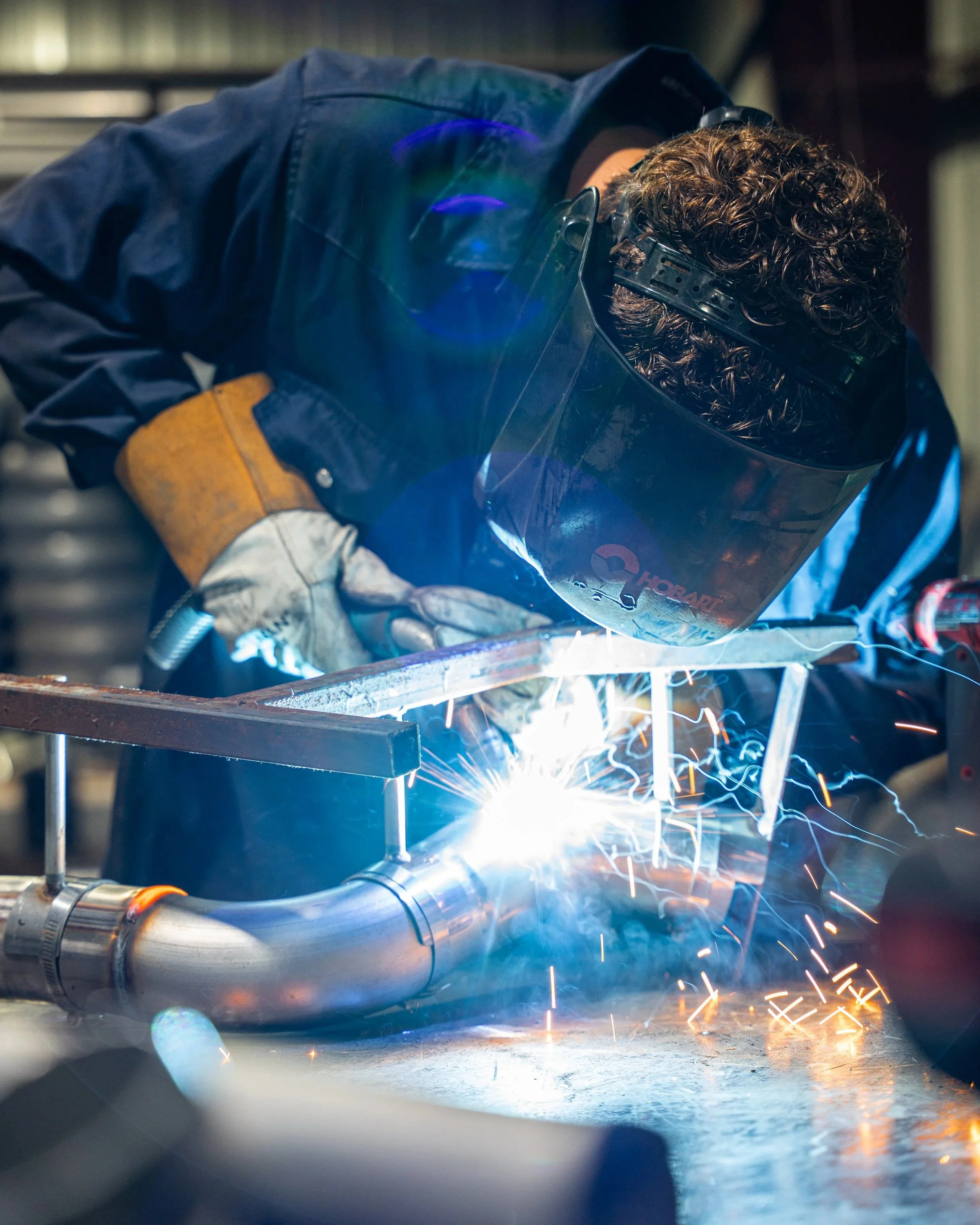 A person welding metal pipes in a workshop, wearing protective gear including a welding helmet and gloves, with sparks flying from the welding process.