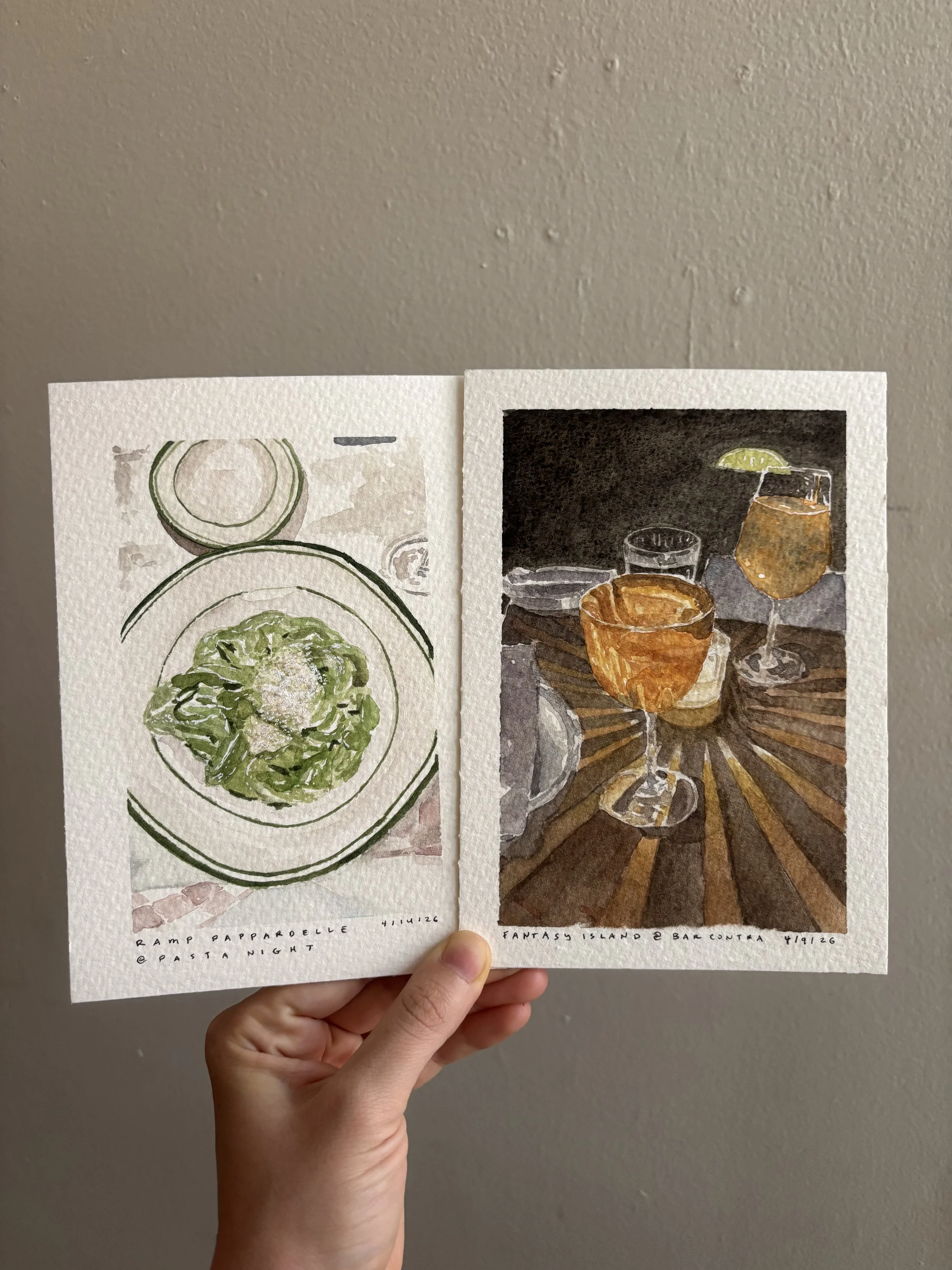 Two watercolor paintings of food and drinks, one featuring a bowl of salad on a plate and the other depicting glasses of cocktails with a lime wedge on a dark background, held by a person's hand against a gray wall.