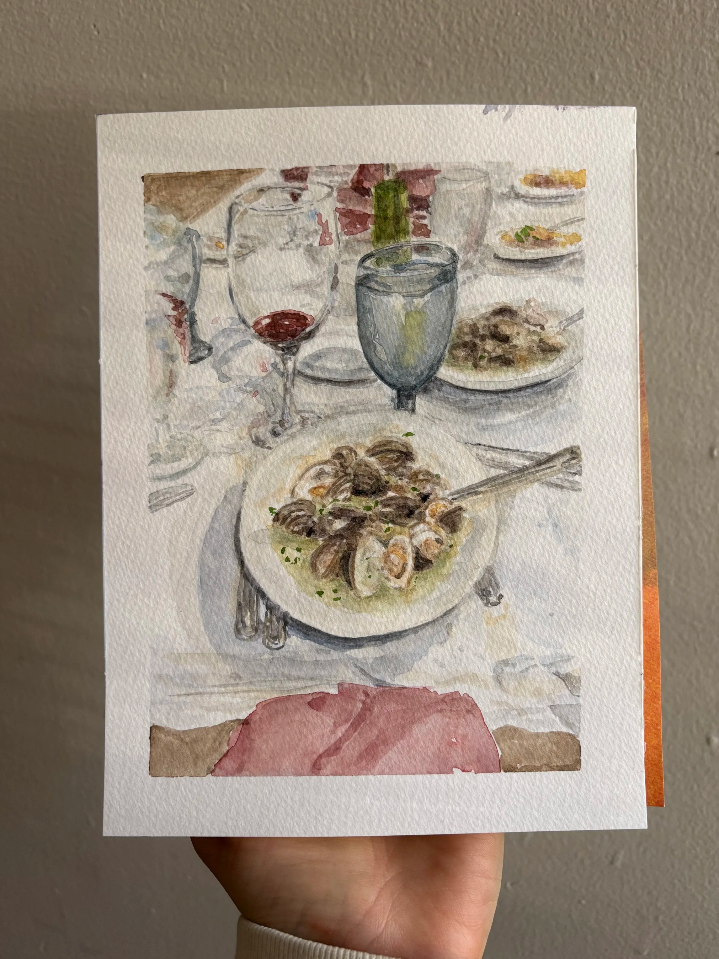 Hand holding a watercolor painting of a dining table with glasses of wine, plates of food, and a bottle, against a plain wall.