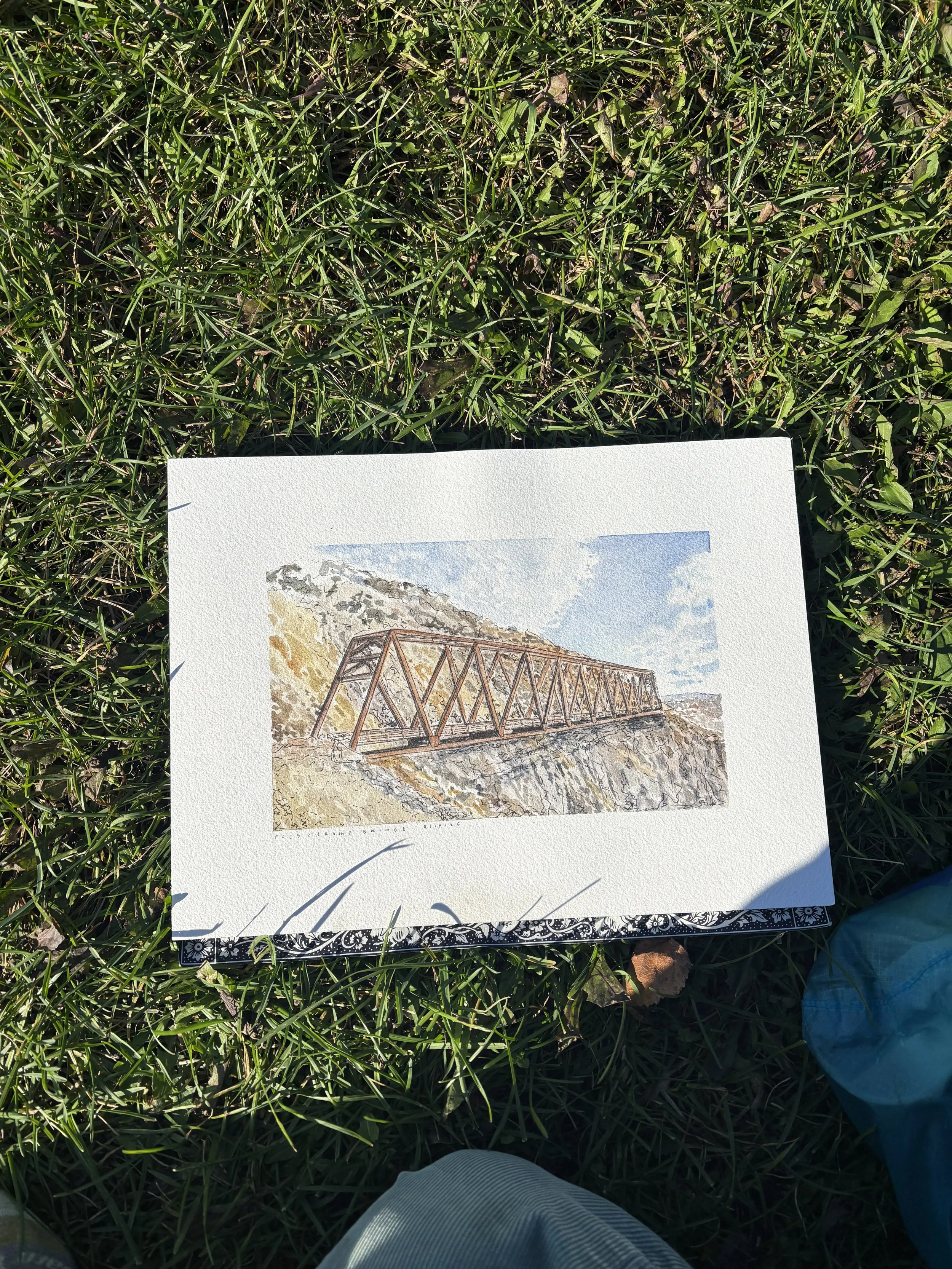 A watercolor painting of a wooden bridge over a rocky landscape with a cloudy sky in the background.