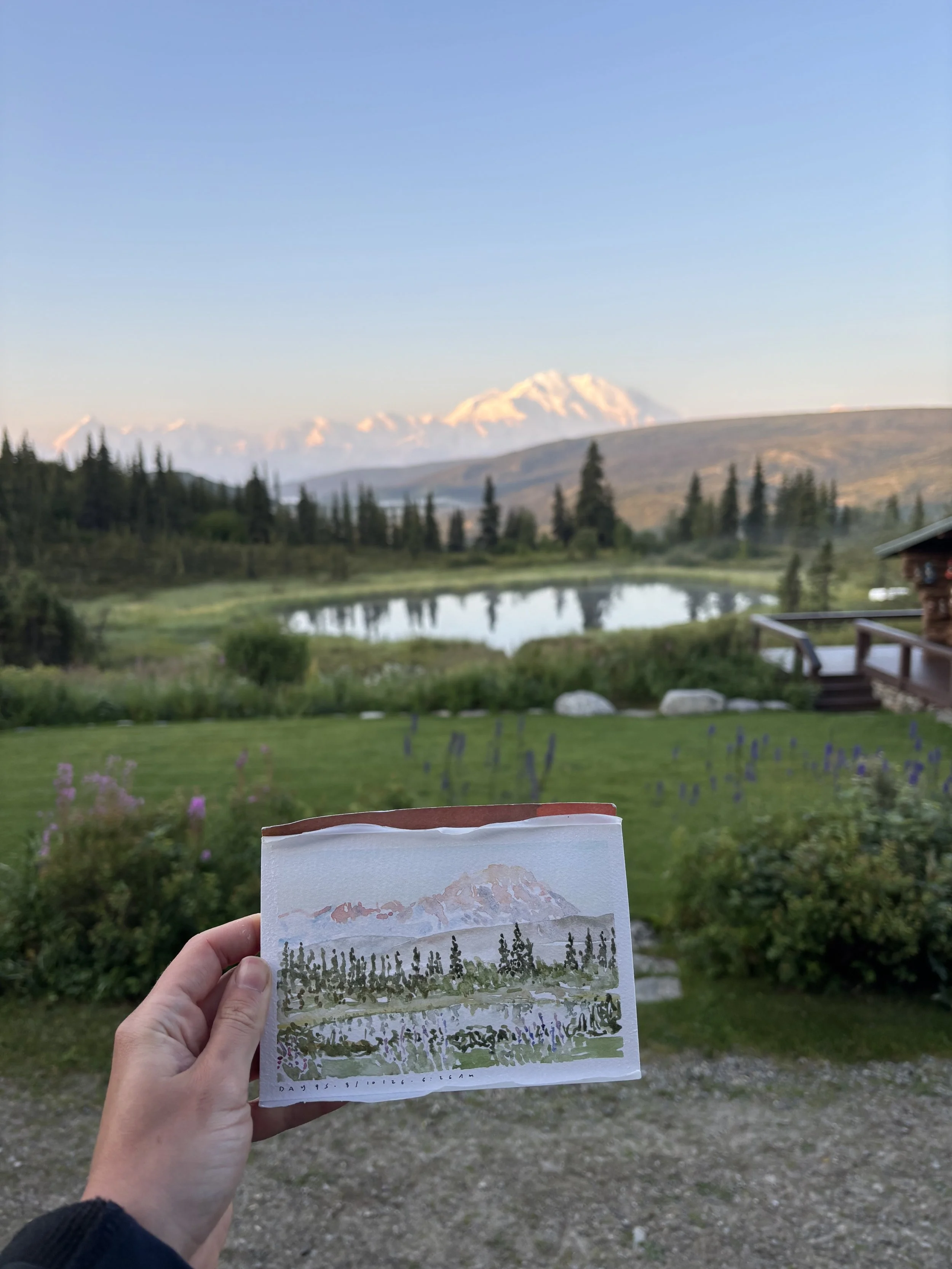 A person holding a small watercolor painting of a mountain landscape in front of a scenic outdoor mountain view with a lake, trees, and distant snow-capped mountains under a clear blue sky.