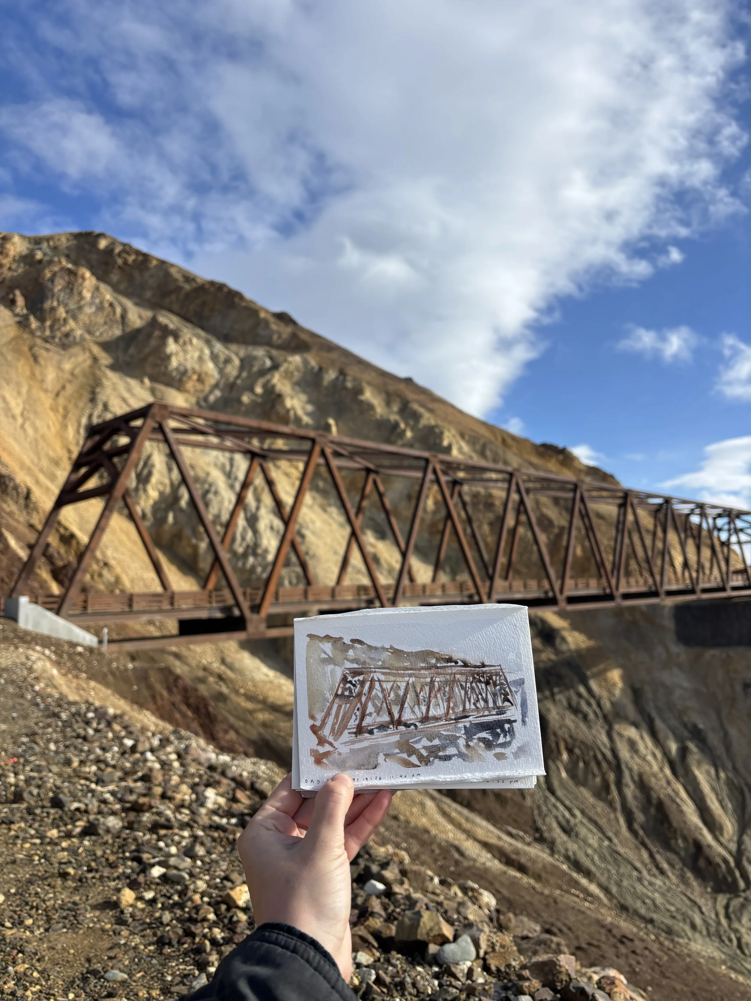 A person holding a small watercolor painting or sketch of a bridge, with a mountain and blue sky with clouds in the background.