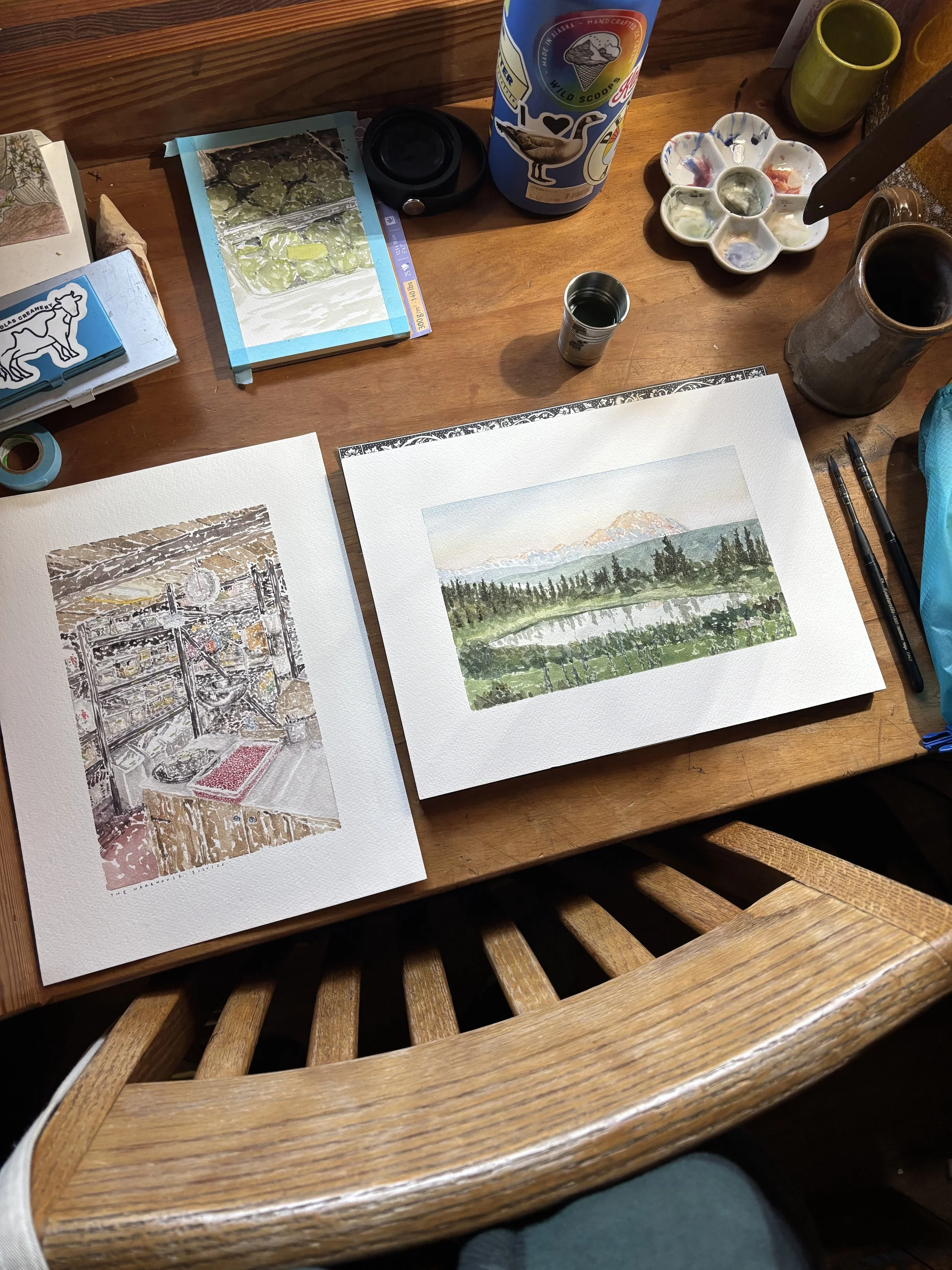 Two watercolor landscape paintings on a wooden table surrounded by art supplies, including brushes, a paint palette, watercolor paints, and cups of water.