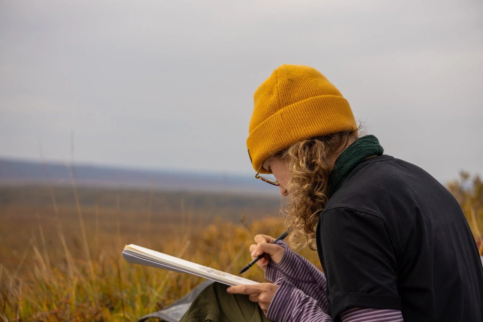Person with curly hair wearing a yellow beanie, glasses, and a black jacket, sitting outdoors and writing in a notebook during autumn.