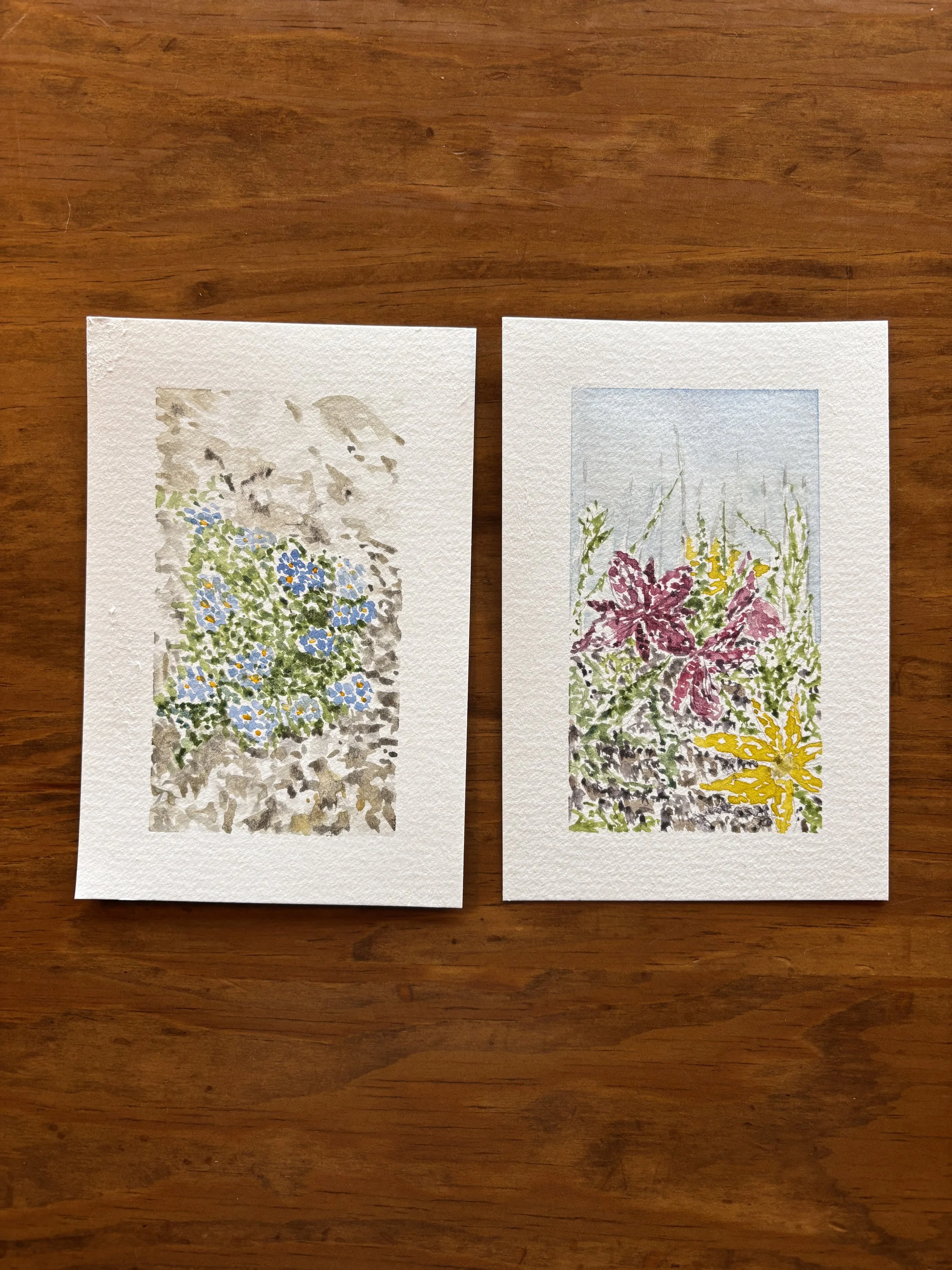 Two pieces of watercolor art depicting colorful floral arrangements on textured paper, placed on a wooden surface.