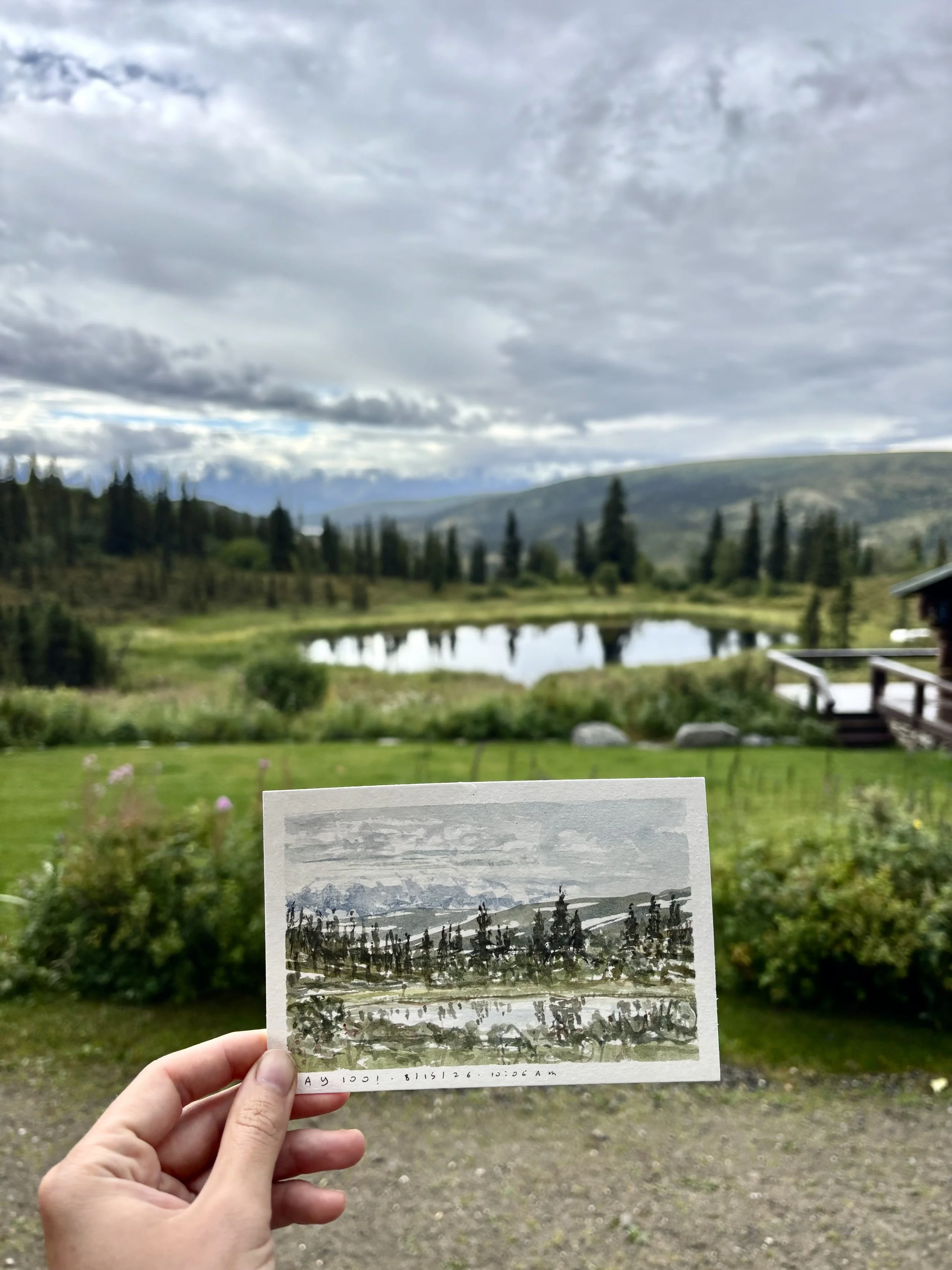 Hand holding a watercolor painting of a landscape with trees, mountains, and a cloudy sky, with a natural landscape and lake in the background.