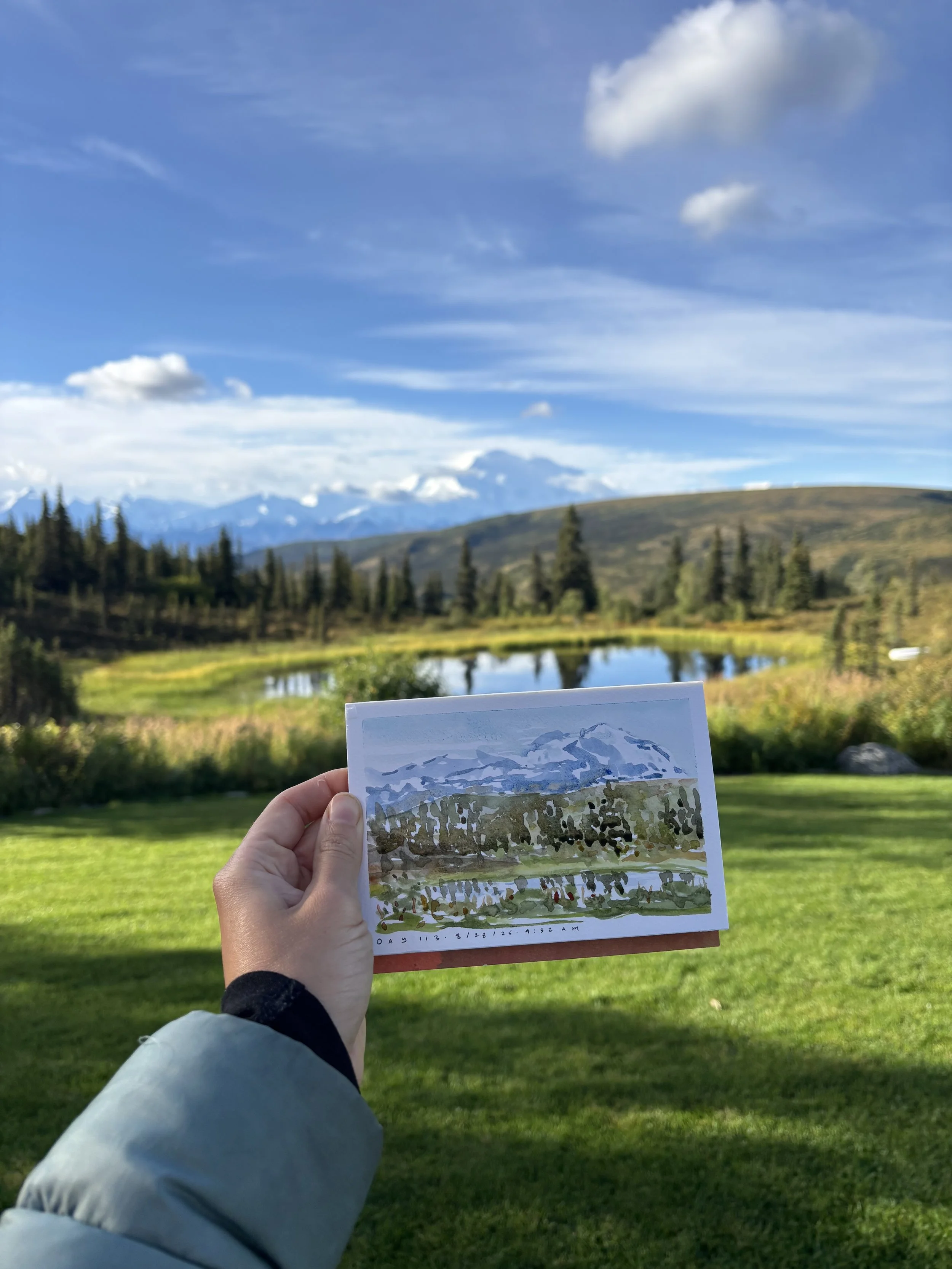 Person holding a sketchbook with a drawing of a mountain landscape in front of a real mountain view, with a lake, trees, and a blue sky with clouds.